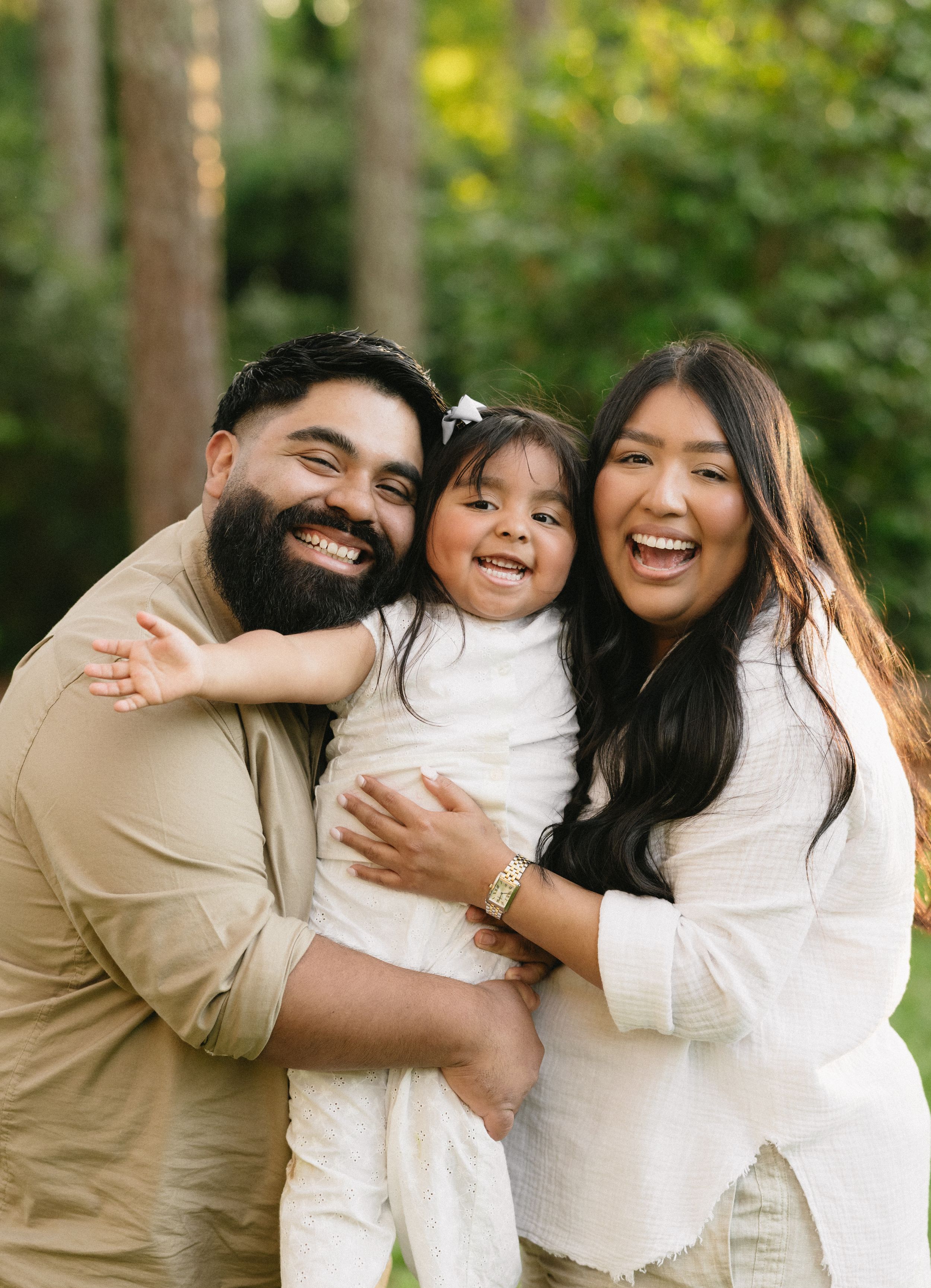 A family shares joyful embraces and laughter while wearing coordinated white and beige outfits outdoors.