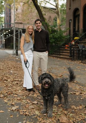 Couple walking black dogs on leaf-covered path near historic buildings.