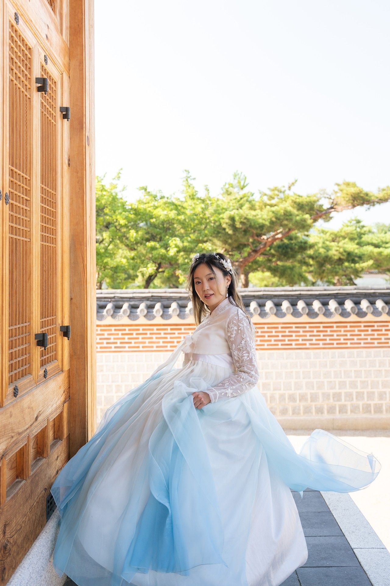 A traditional Korean hanbok dress with flowing white and blue fabric is photographed in a palace courtyard setting.