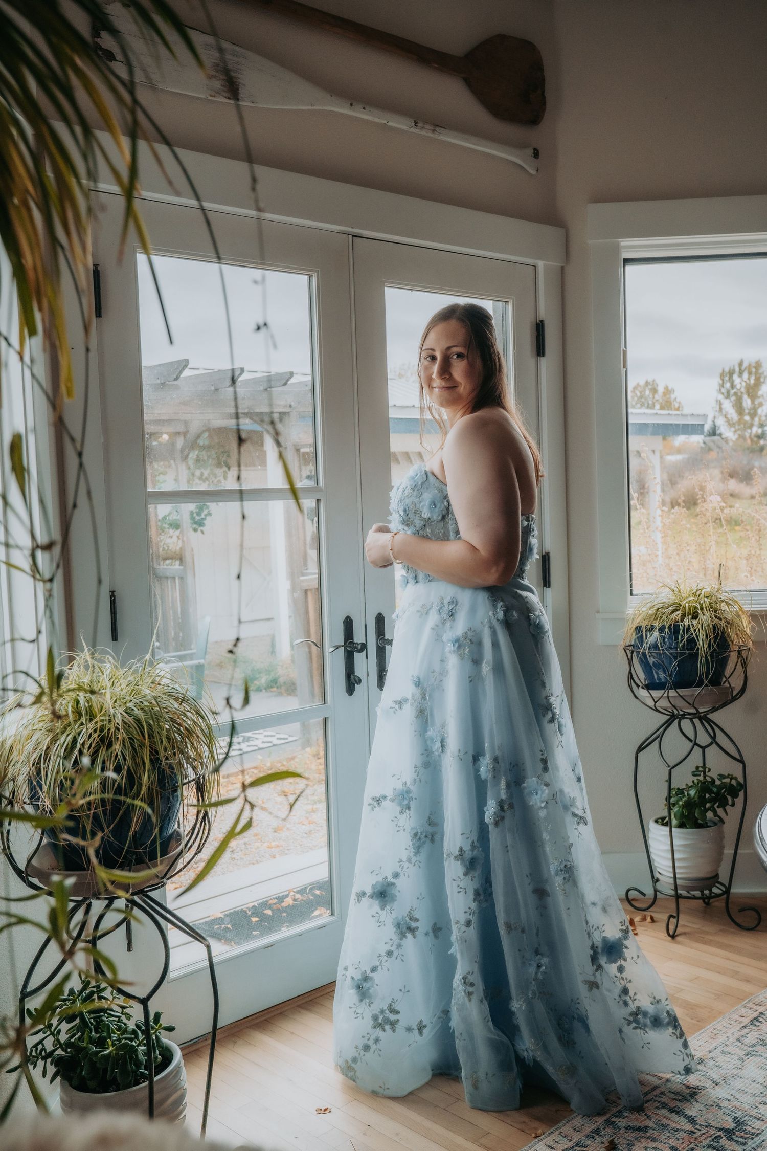 A person in a flowing light blue floral gown stands by large windows in a sunlit room with hanging plants.