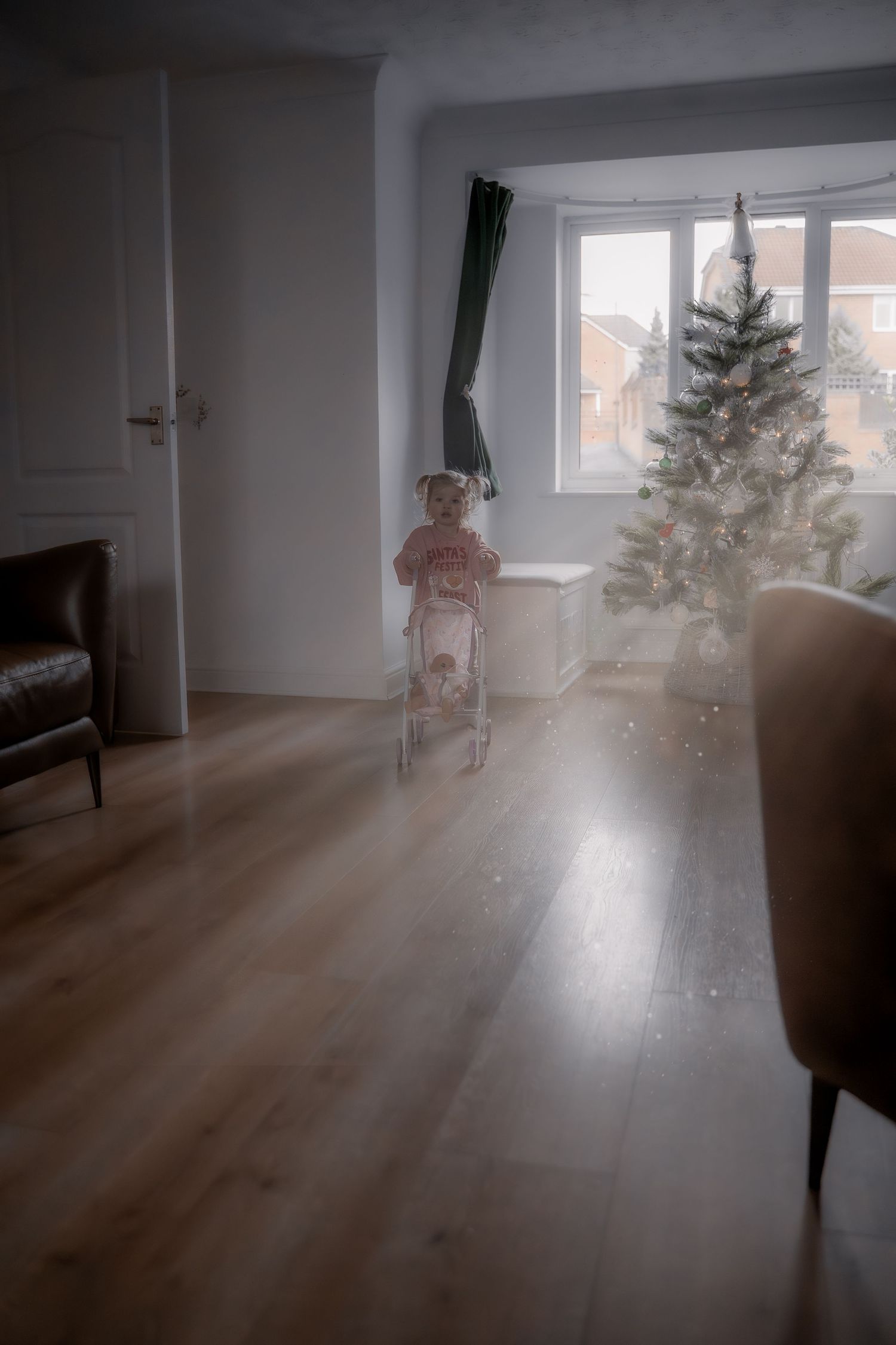 A dreamy interior scene shows a small figure by a Christmas tree near a window in a room with hardwood floors.