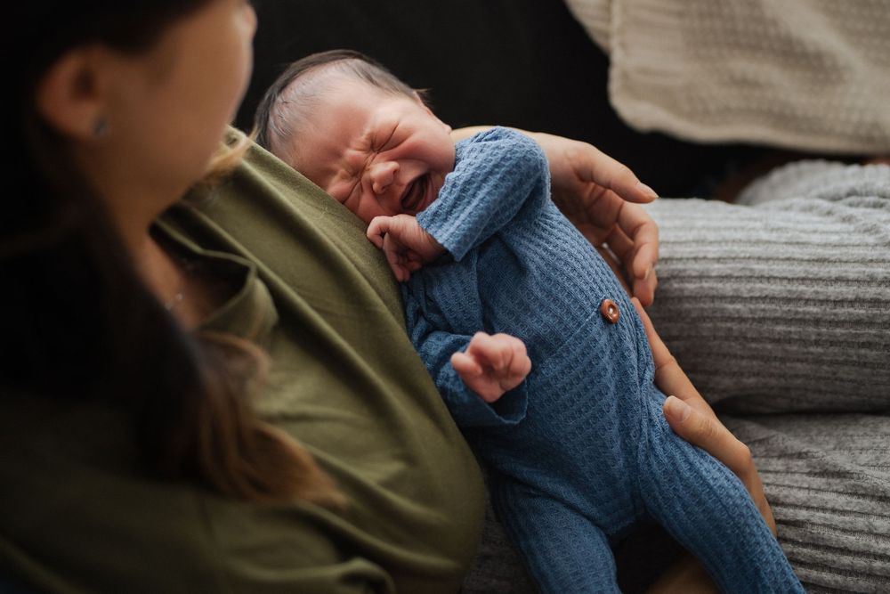 Newborn baby in blue knit outfit yawns while being cradled on cozy couch.