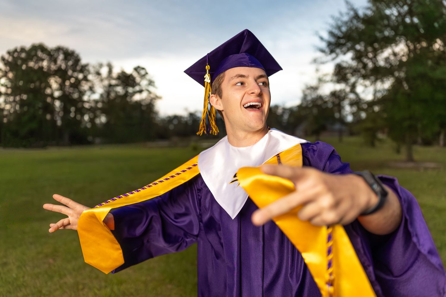 Graduate in purple cap and gown celebrating with arms outstretched outdoors.