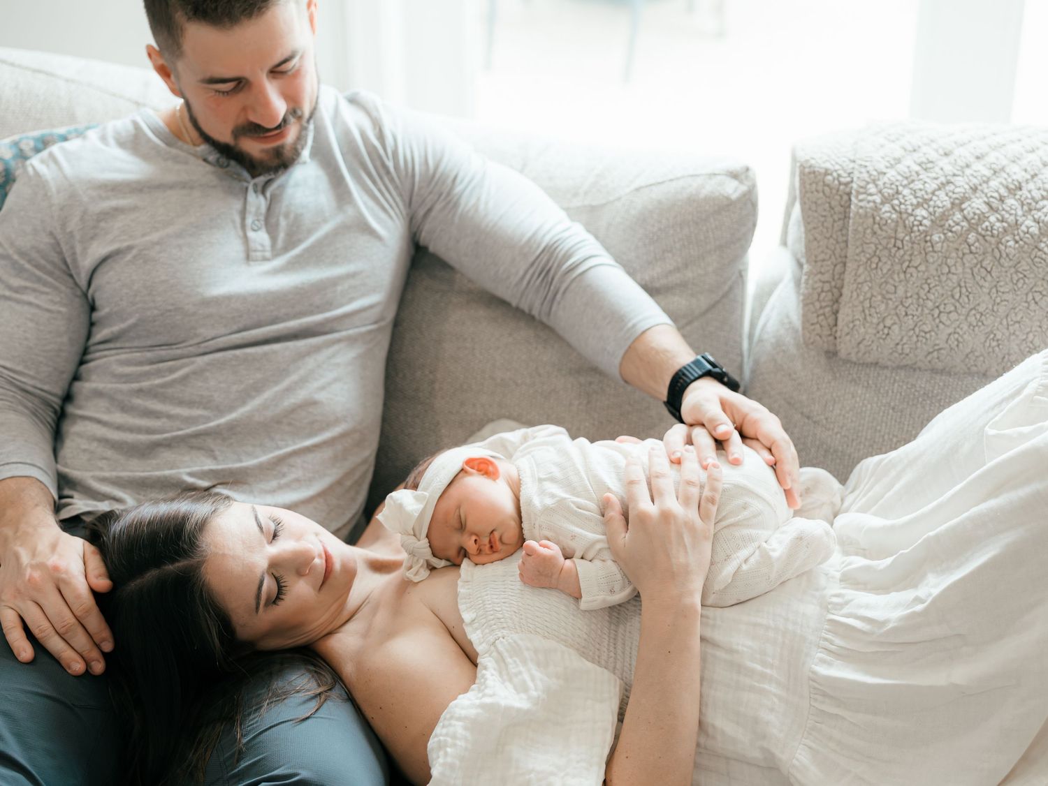A new mother holds her newborn while laying on her husbands leg snuggled on the couch.