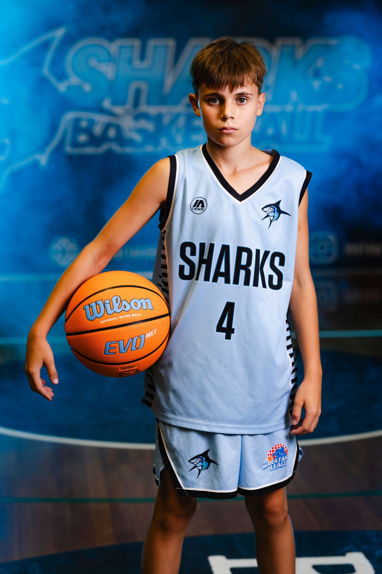 Young basketball player in white Sharks uniform holds Wilson basketball against blue backdrop.