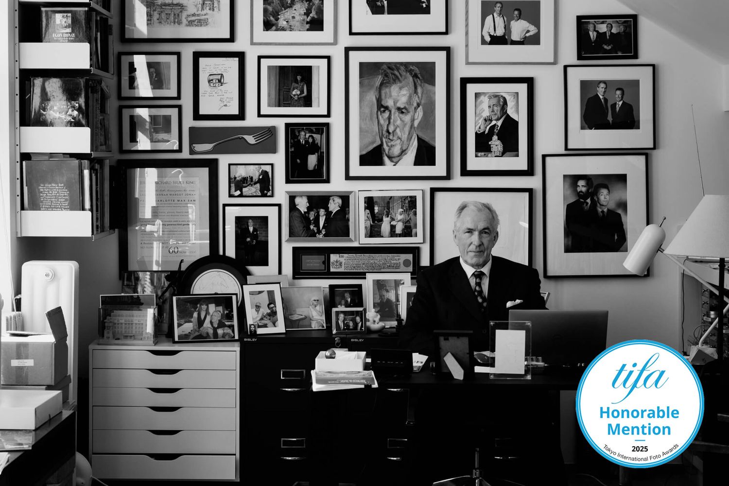 Black and white photograph of a wall filled with framed portraits and photographs in a gallery-style arrangement.