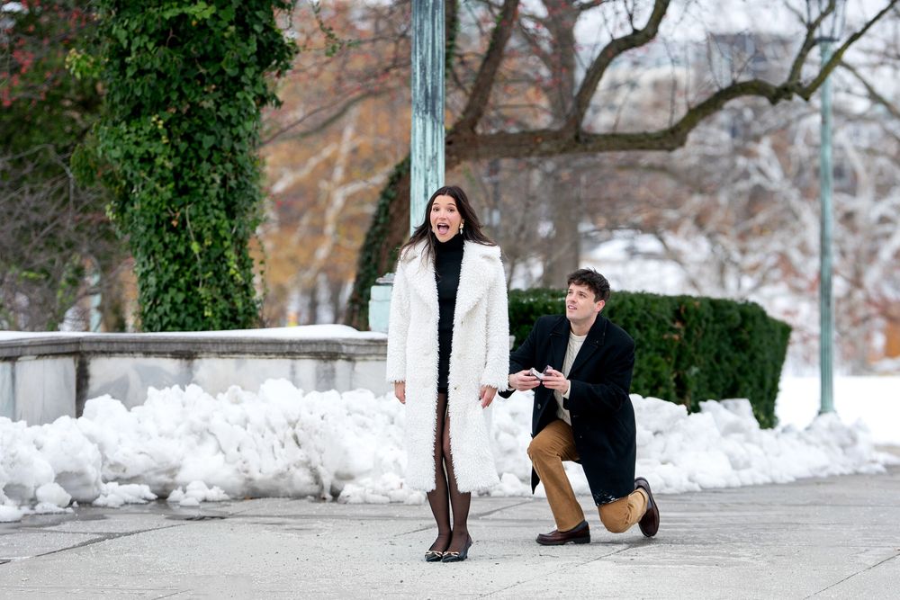 Man proposes on one knee to surprised woman in white coat in snowy winter park.