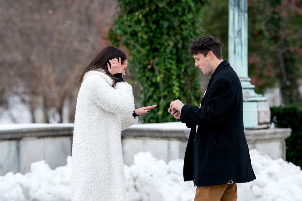 Man opening ring box while emotional woman covers her face during snowy winter marriage proposal outdoors.