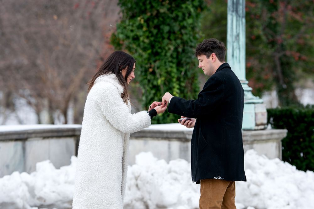 Man placing engagement ring on womans finger during romantic winter proposal in snowy park setting.