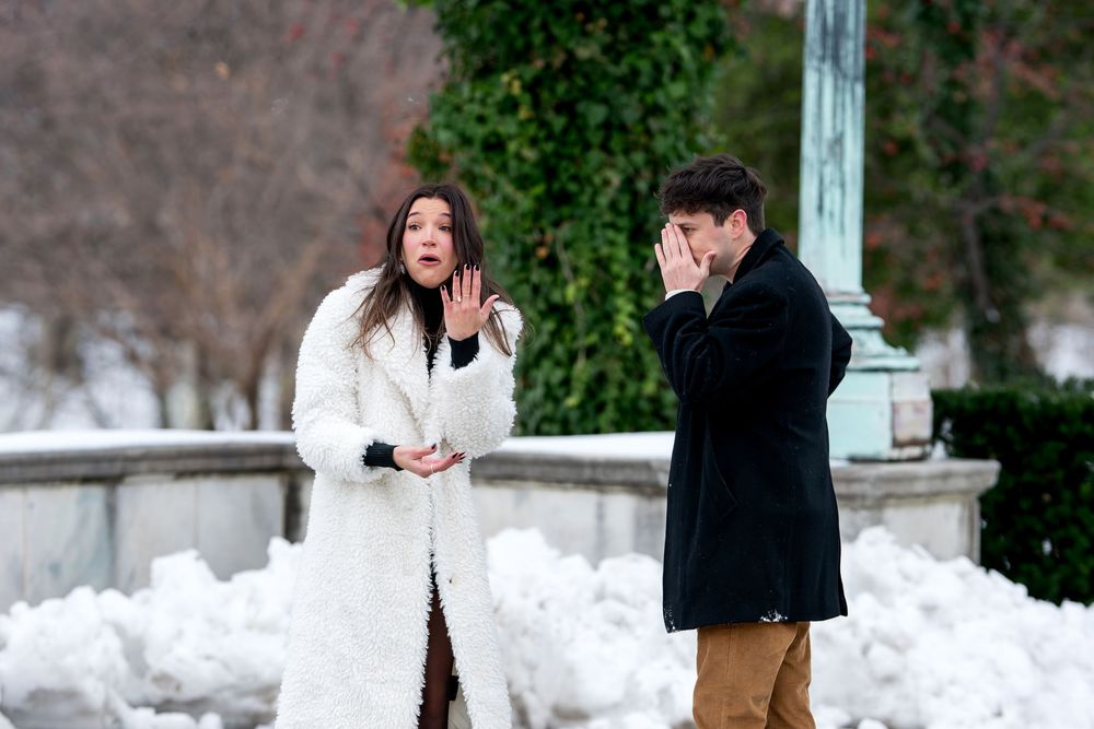 Surprised woman gestures while man covers face with emotion during a winter marriage proposal outdoors.