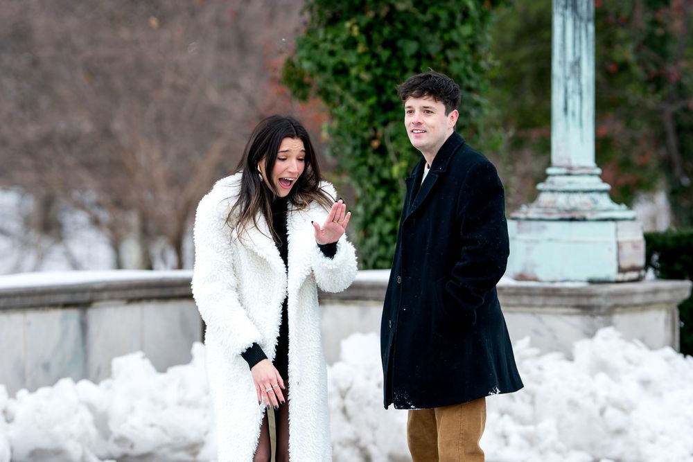 Excited woman admires engagement ring while smiling man stands beside her in snowy winter park.
