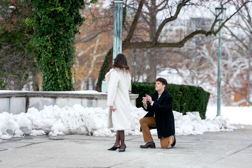 Man on one knee proposing to woman in elegant white coat in snow-covered park.