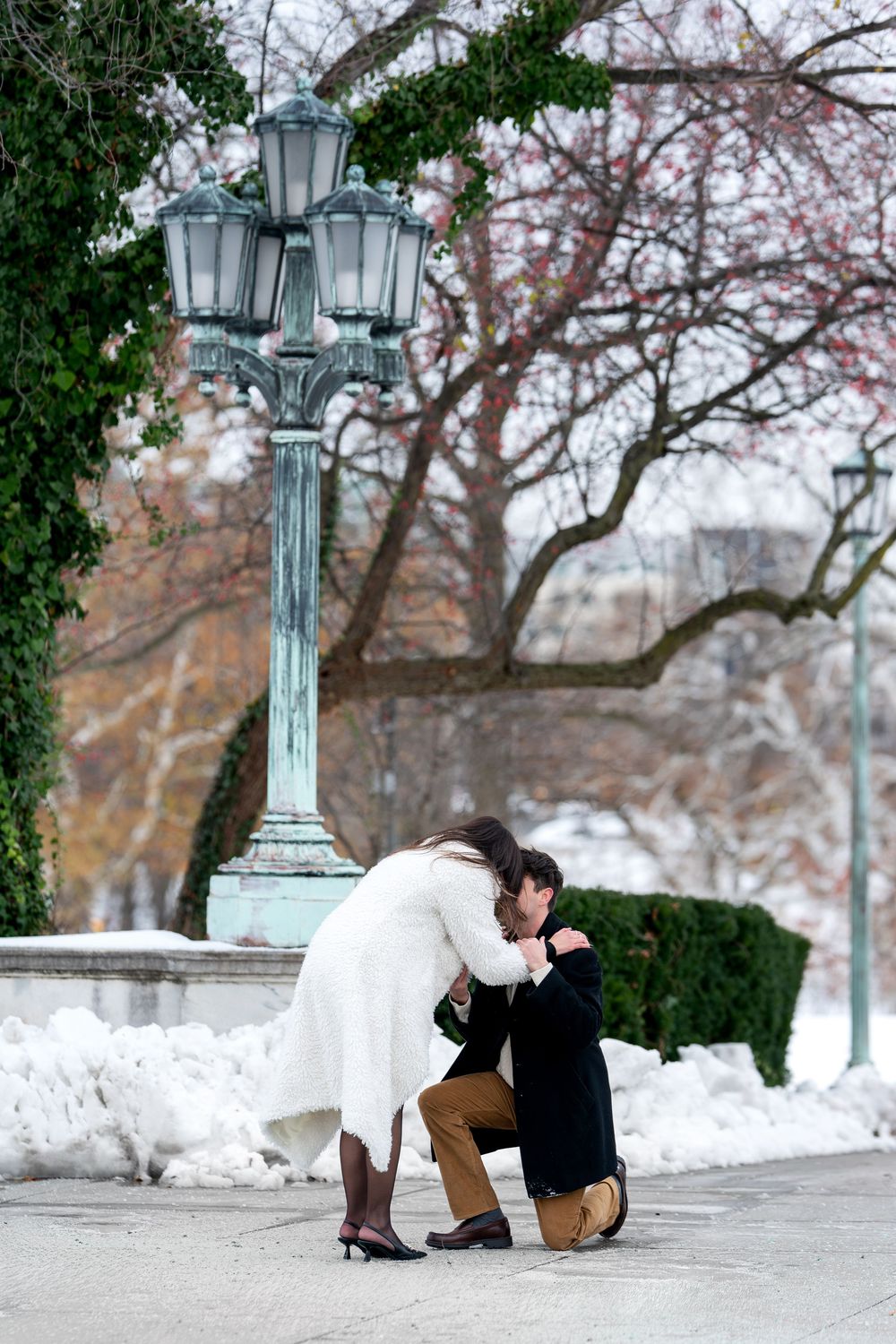 Woman leans down to embrace man during heartfelt winter marriage proposal in park.