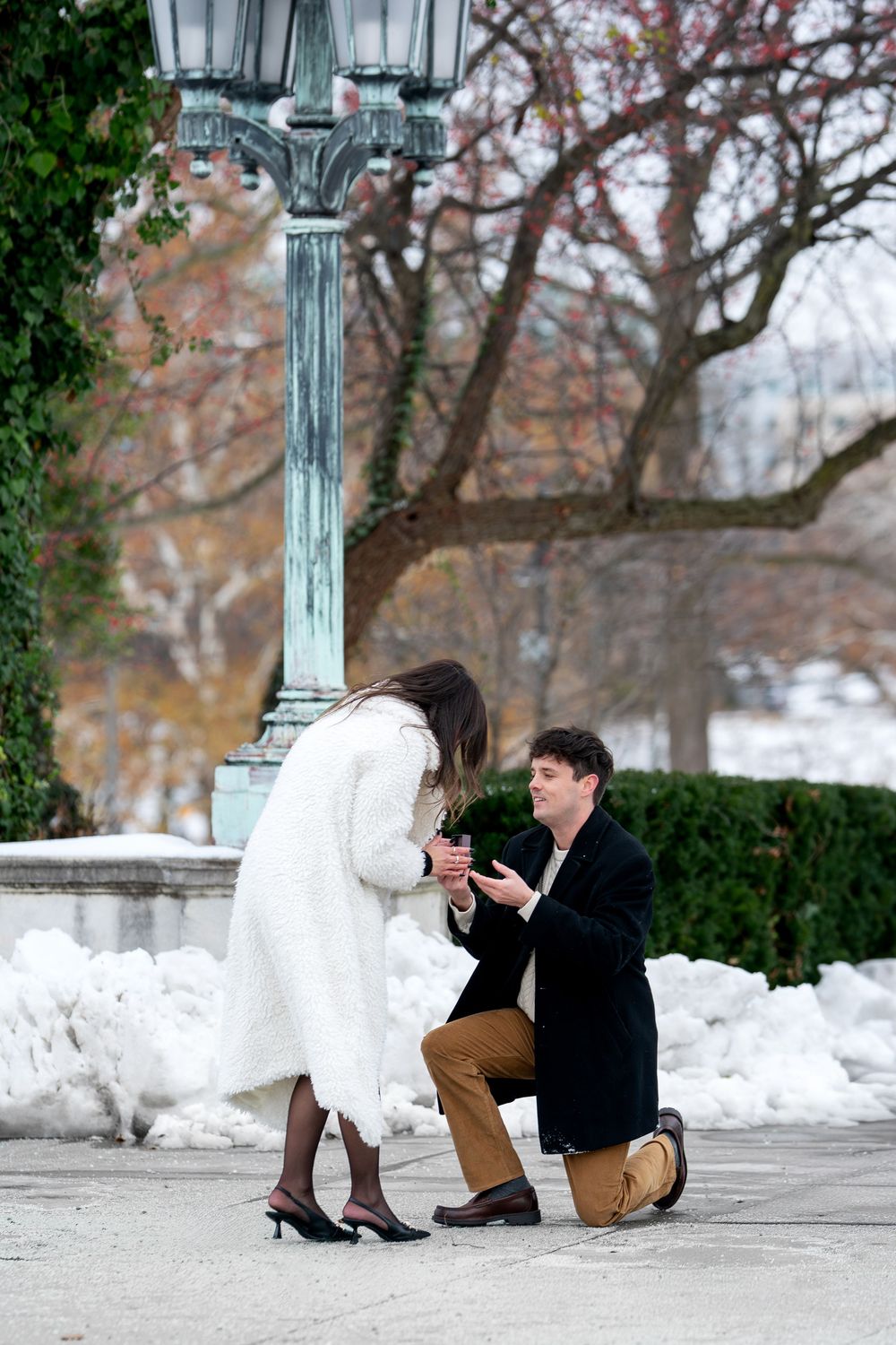 Man presents engagement ring while kneeling before woman in white coat in snowy park.