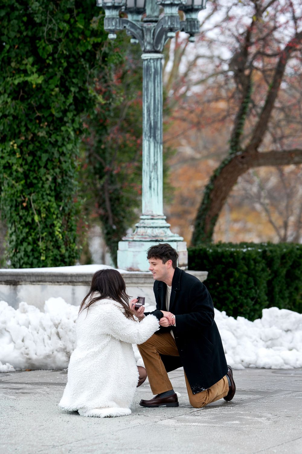 Couple examines engagement ring together kneeling in snowy winter park setting.