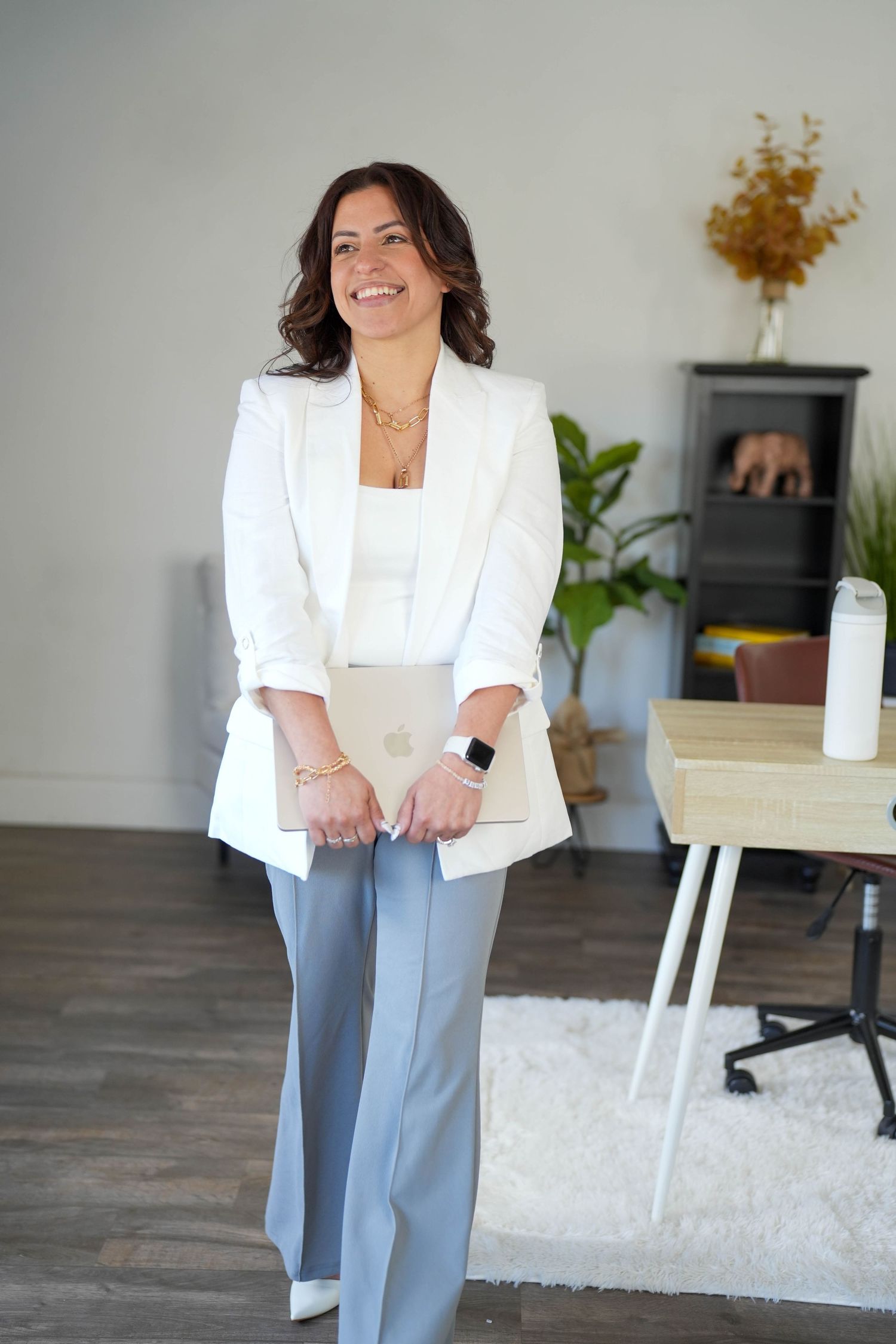 Professional woman in white blazer and gray pants holding a laptop, smiling in a modern office space.