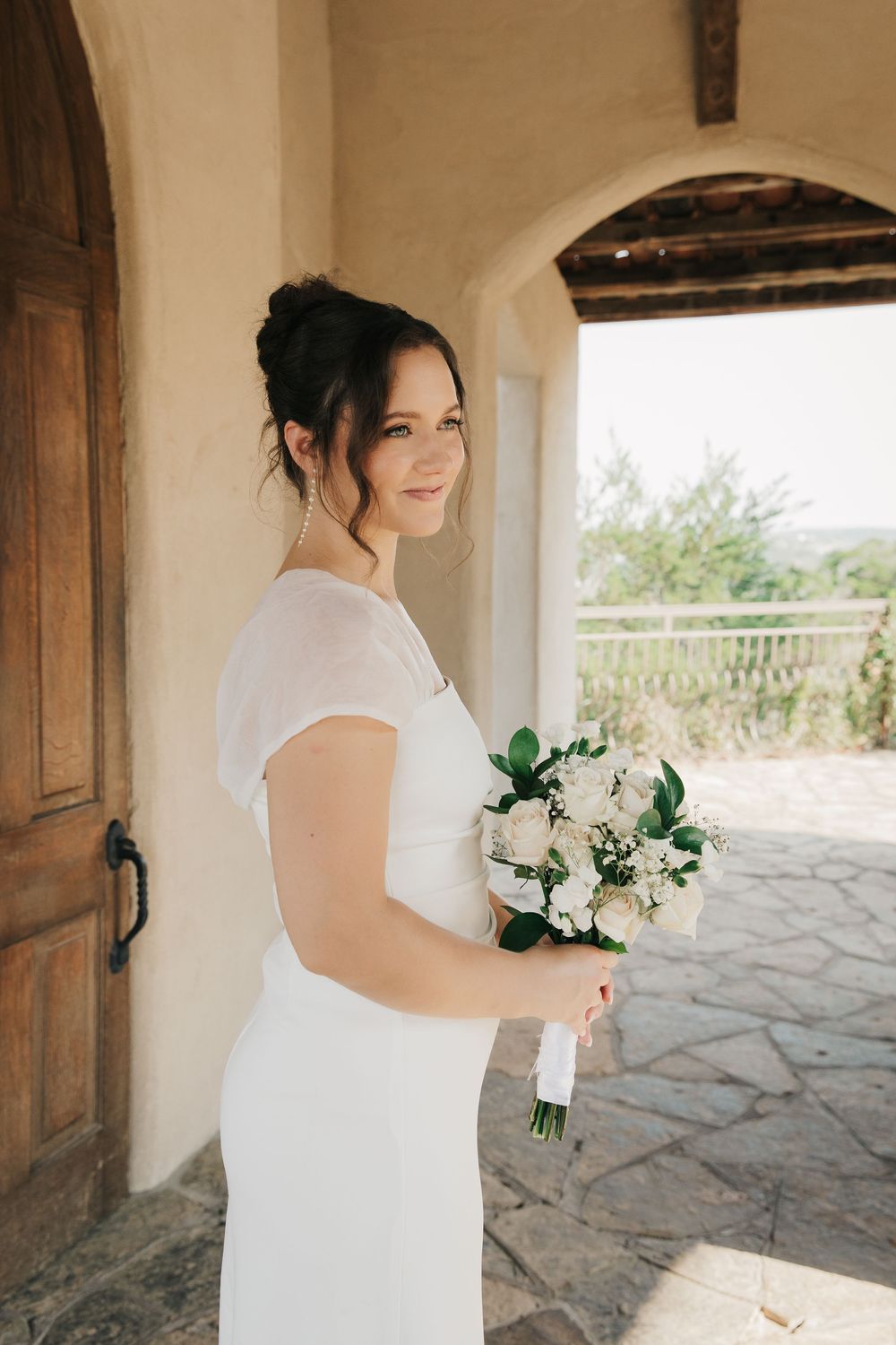 A bride in a white dress holds a bouquet of white flowers while standing in an arched doorway.