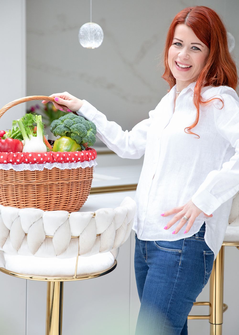 Woman in white shirt and jeans holding various food items and cake on marble countertop in bright kitchen setting.