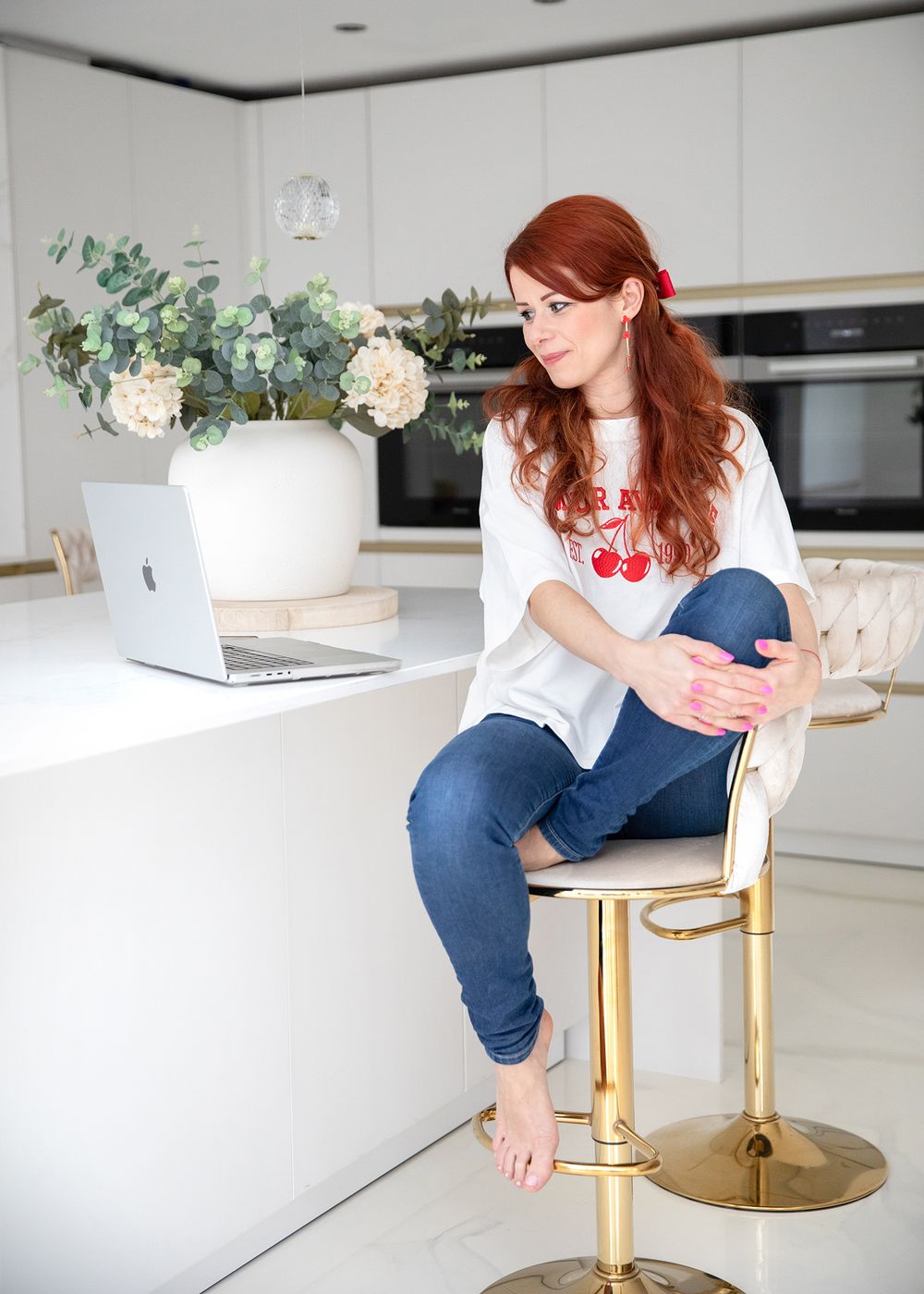 Woman in white top and jeans sits on gold barstool at kitchen counter with laptop and floral arrangement.