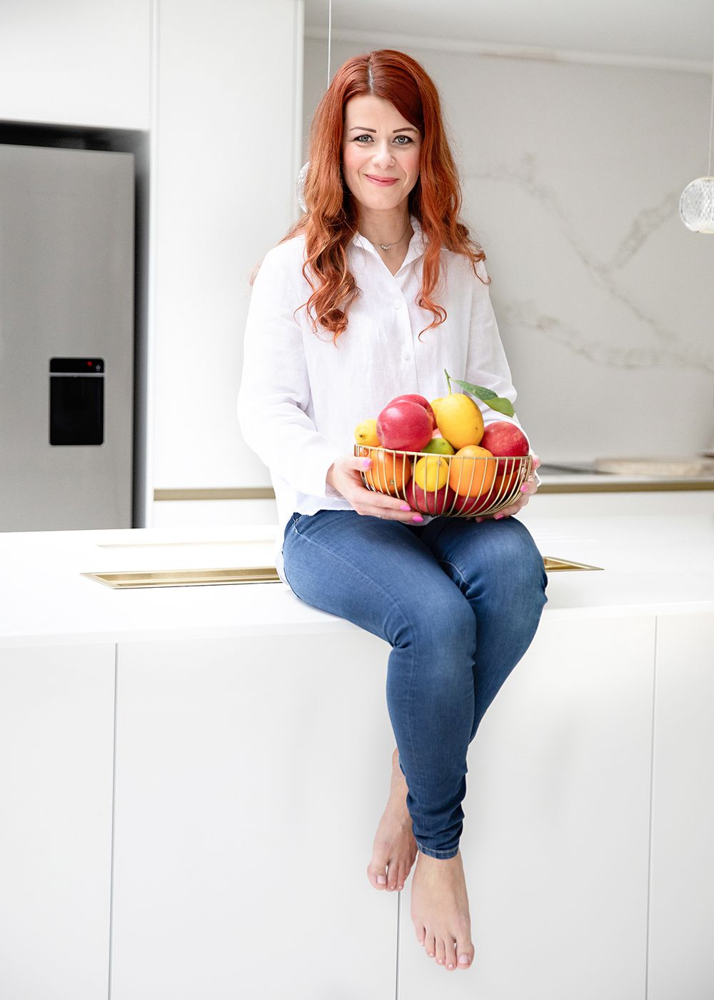 Woman in white top and jeans sits on white counter holding colorful citrus fruits in modern kitchen setting.