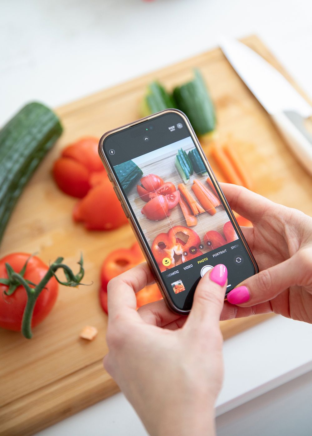 Hands holding a smartphone taking photos of fresh tomatoes and vegetables on a wooden cutting board.