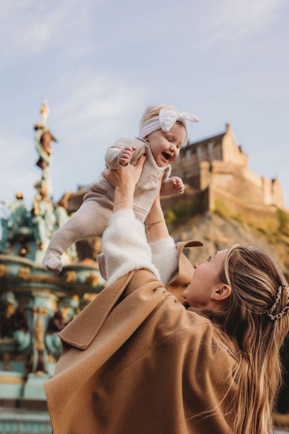 A parent in a tan coat lifts a smiling baby wearing white against an Edinburgh castle backdrop on a cloudy day.