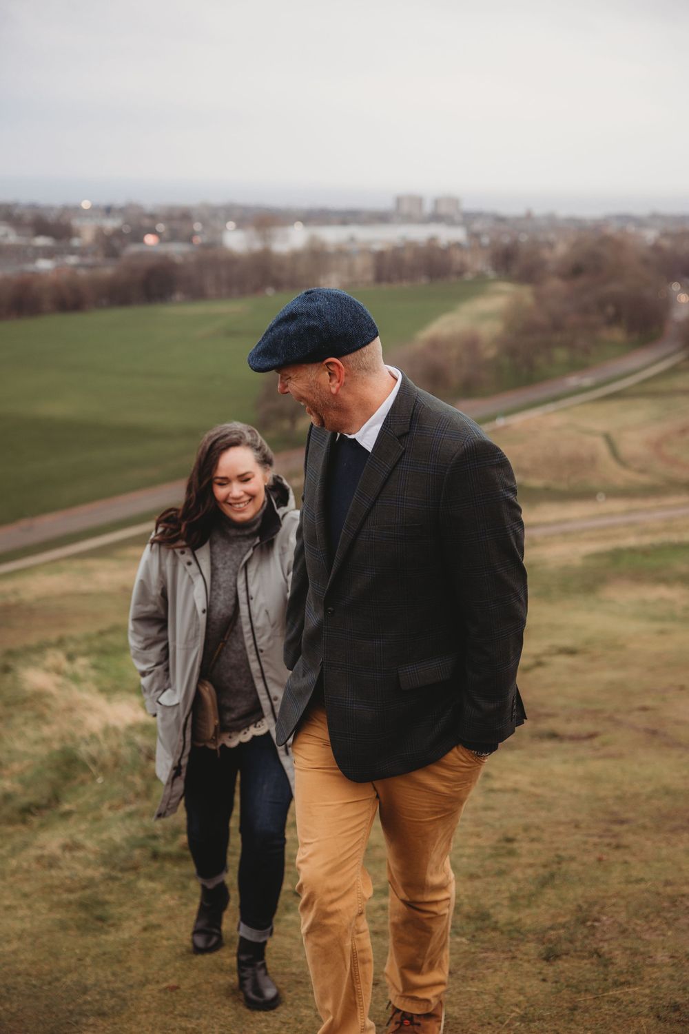 A couple shares a joyful moment together on a hilltop overlooking an urban landscape on an overcast day.