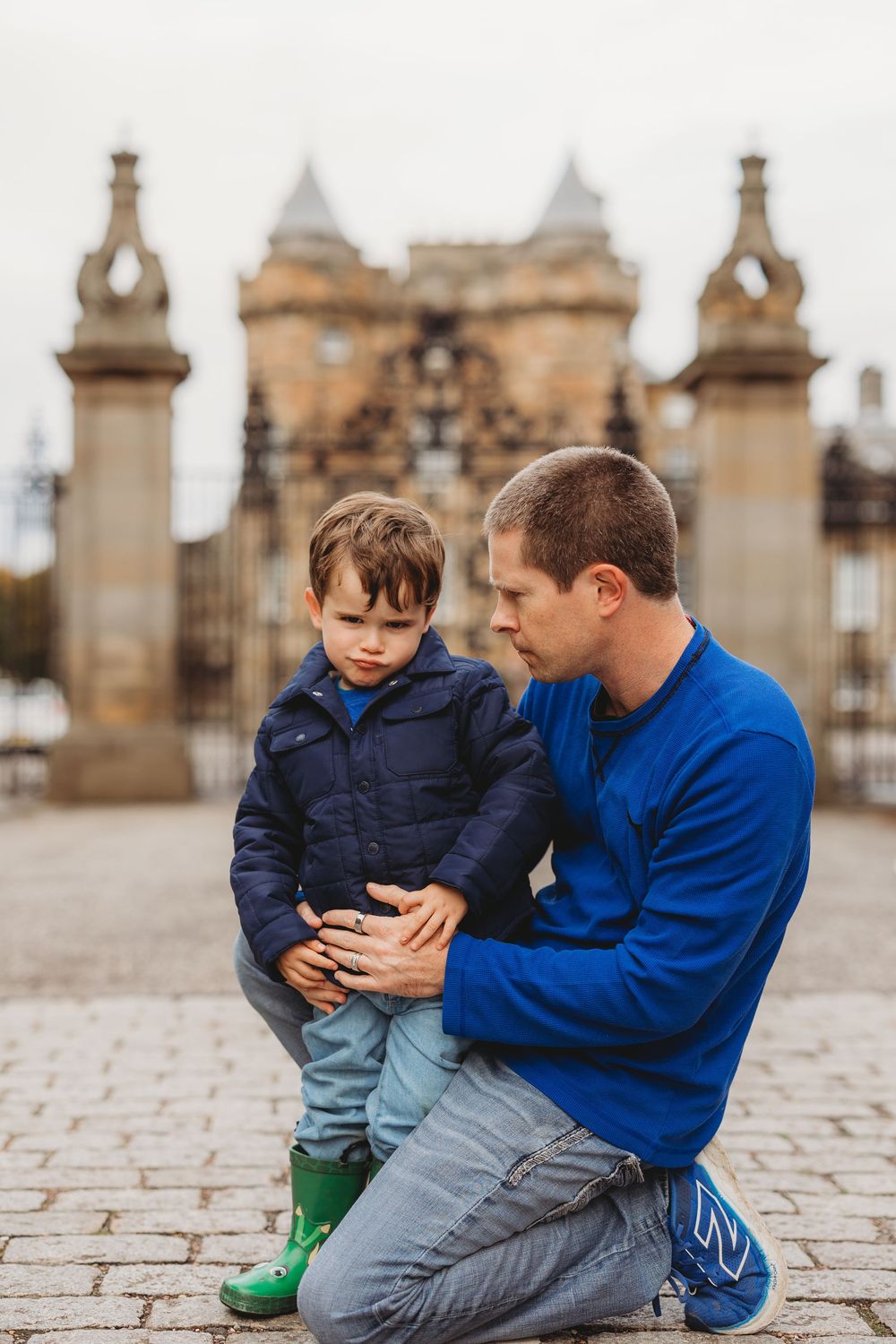A parent and child share a tender moment on cobblestones in front of an ornate castle gate.