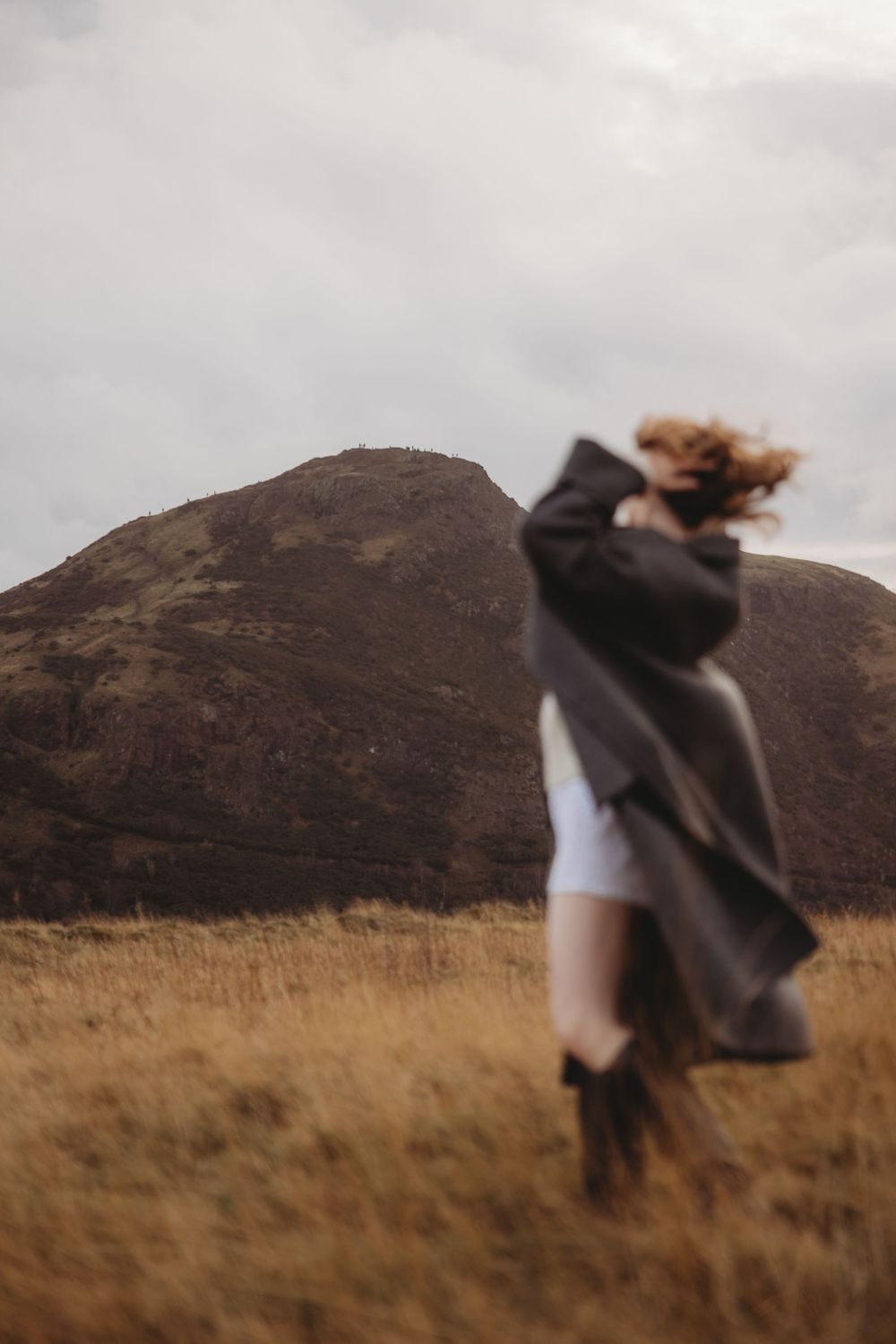 A person in a dark coat dances in a windswept field with mountains in the background at golden hour.