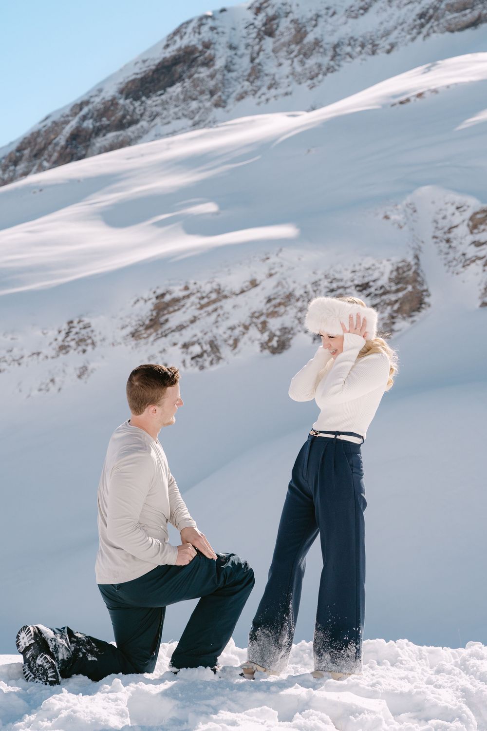 Two people sitting in deep snow against a mountainous winter landscape at sunset.