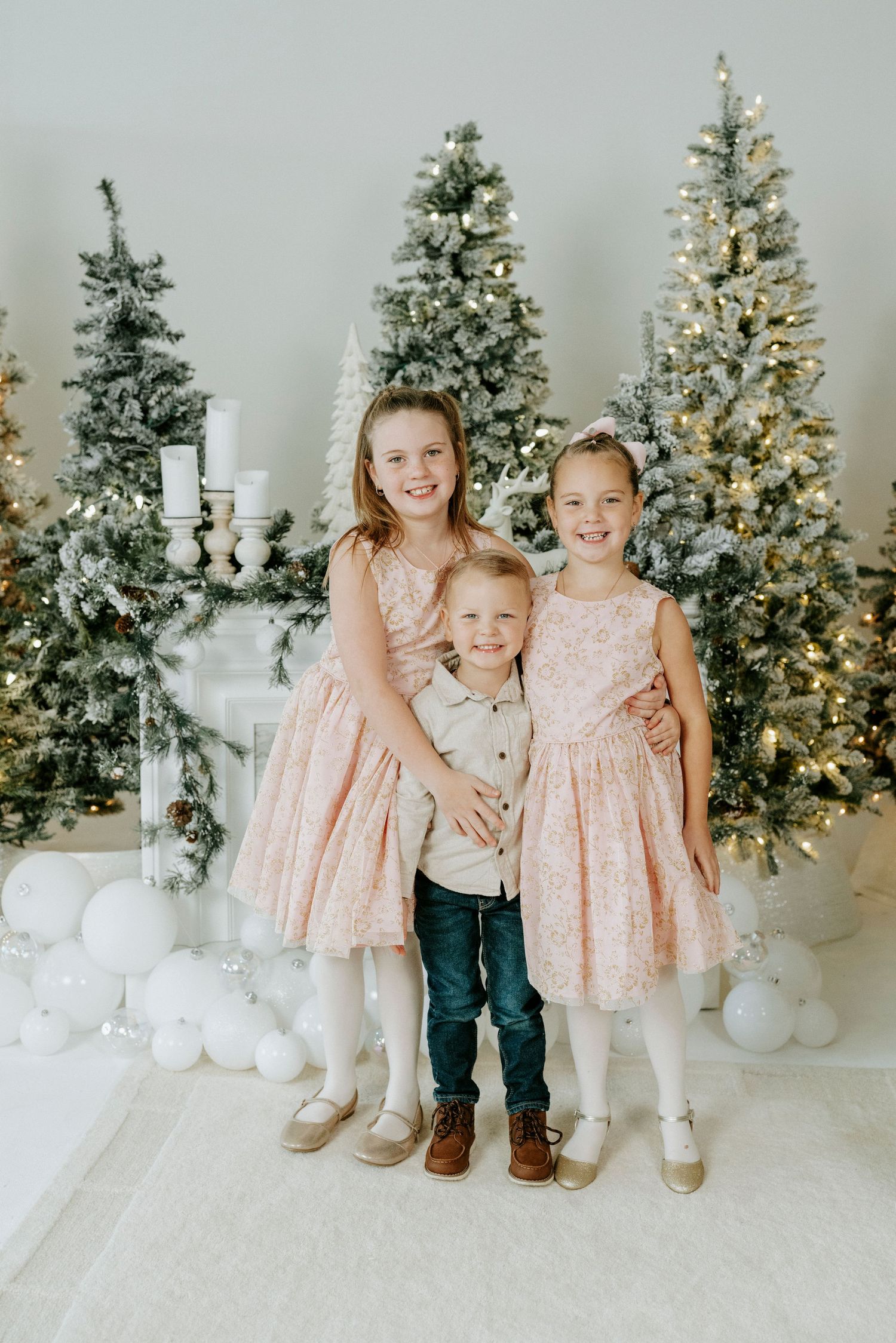 Children pose together in front of decorated Christmas trees and white snowman decorations in a winter studio setting.