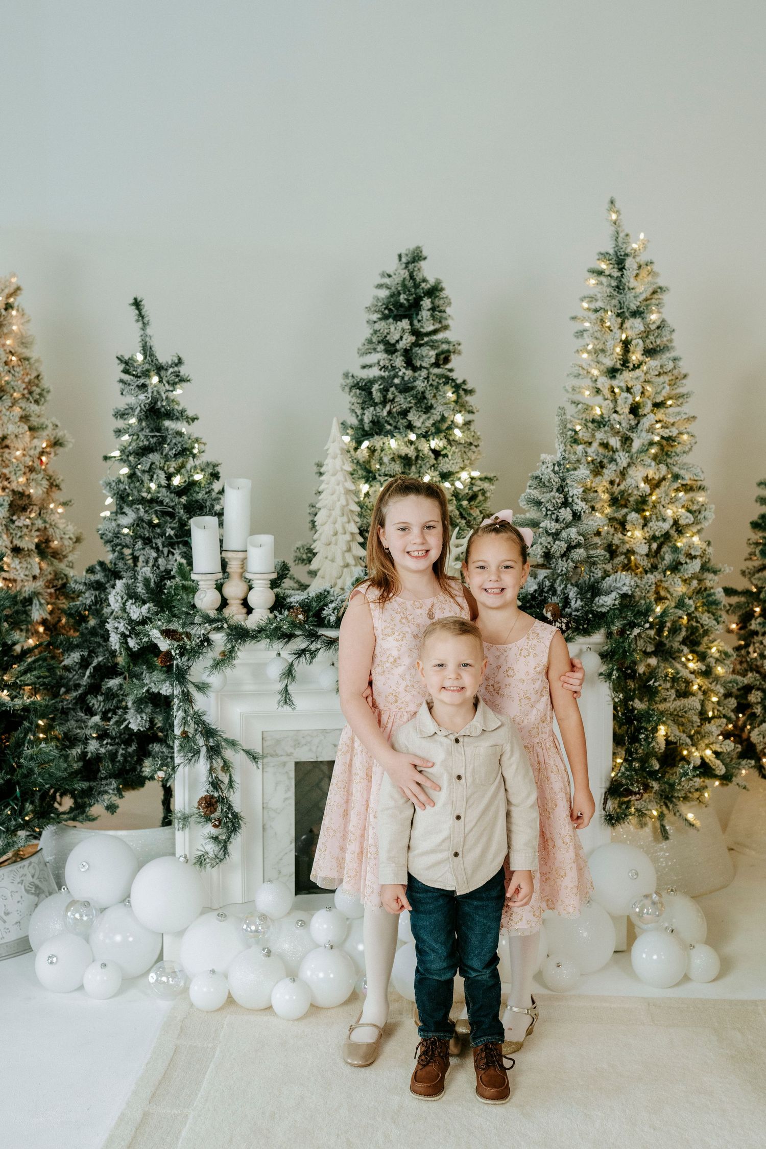 A festive holiday scene with decorated Christmas trees and white ornaments arranged against a light colored wall.