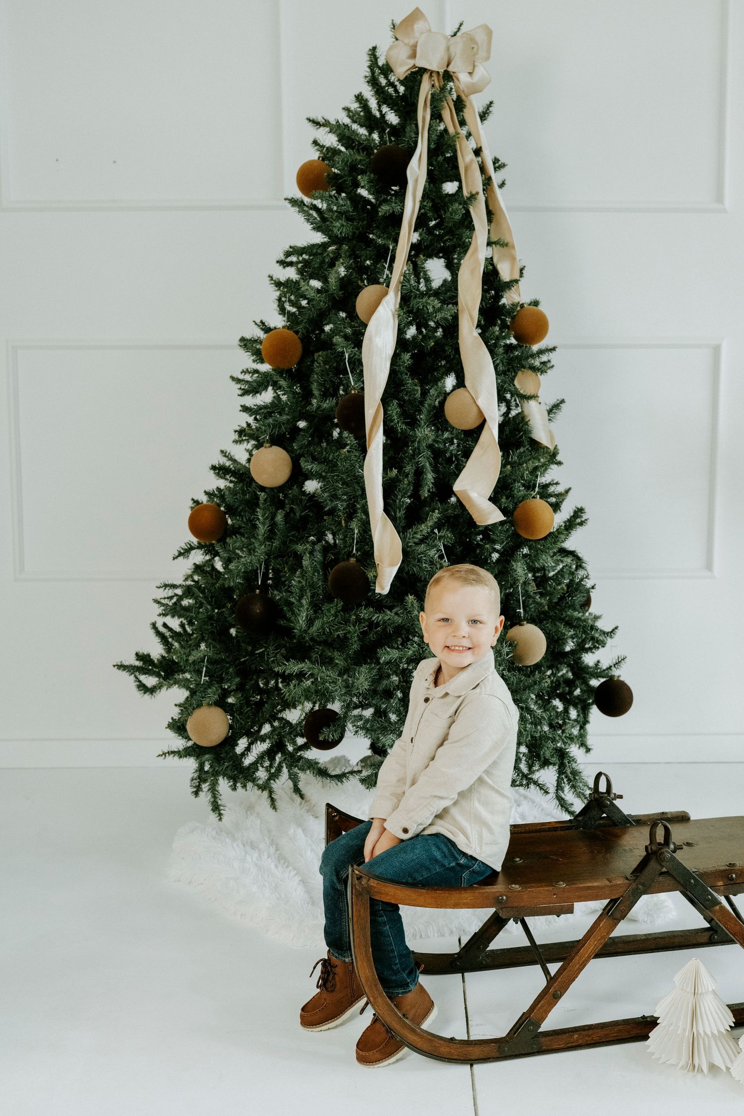 A vintage wooden sled decorated with ribbons sits in front of an evergreen Christmas tree with ornaments.