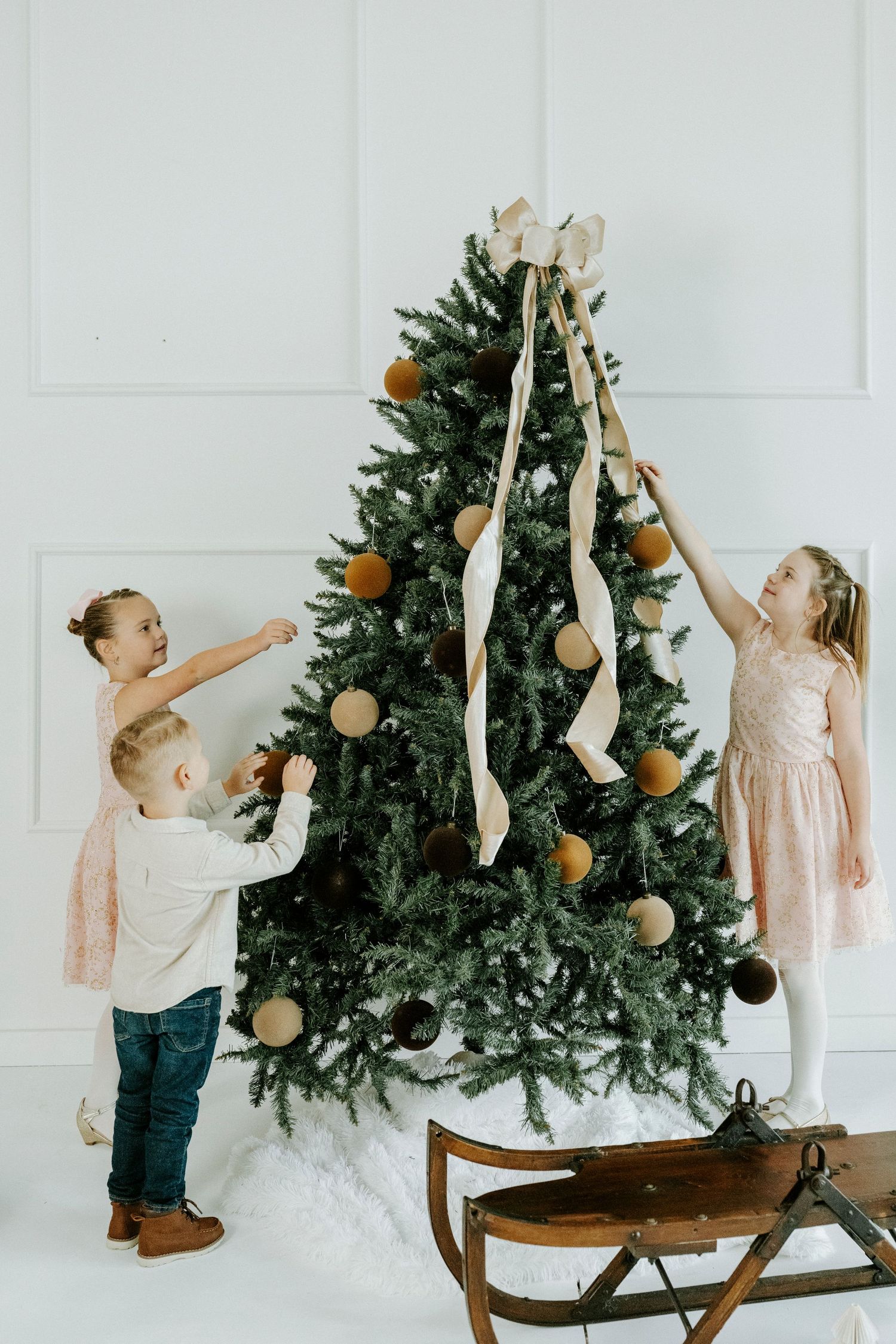 A child in a pink dress and jeans reaches up to decorate a Christmas tree set on a wooden table.