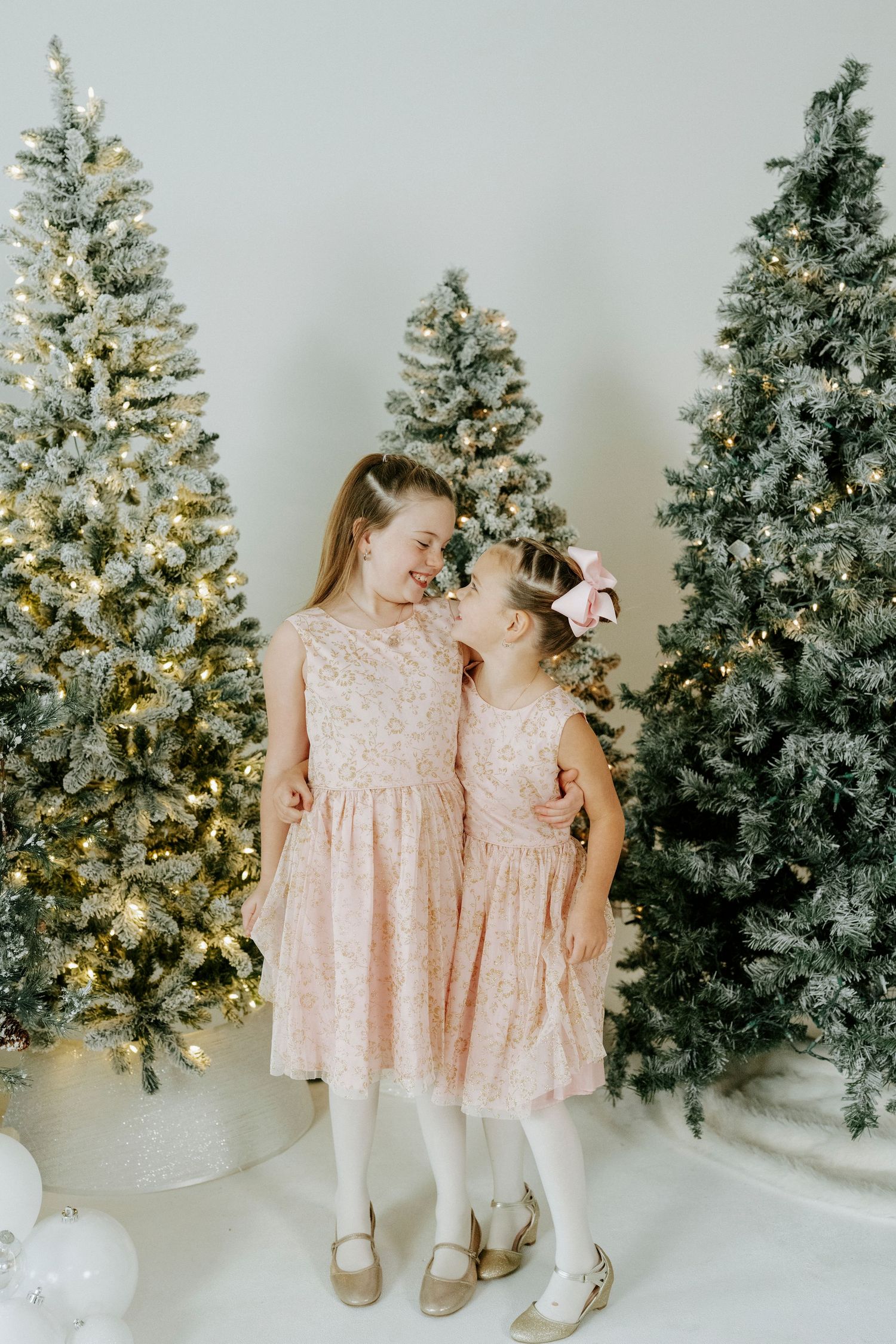 Two children in matching pink dresses sit together near decorated Christmas trees in a winter wonderland setting.