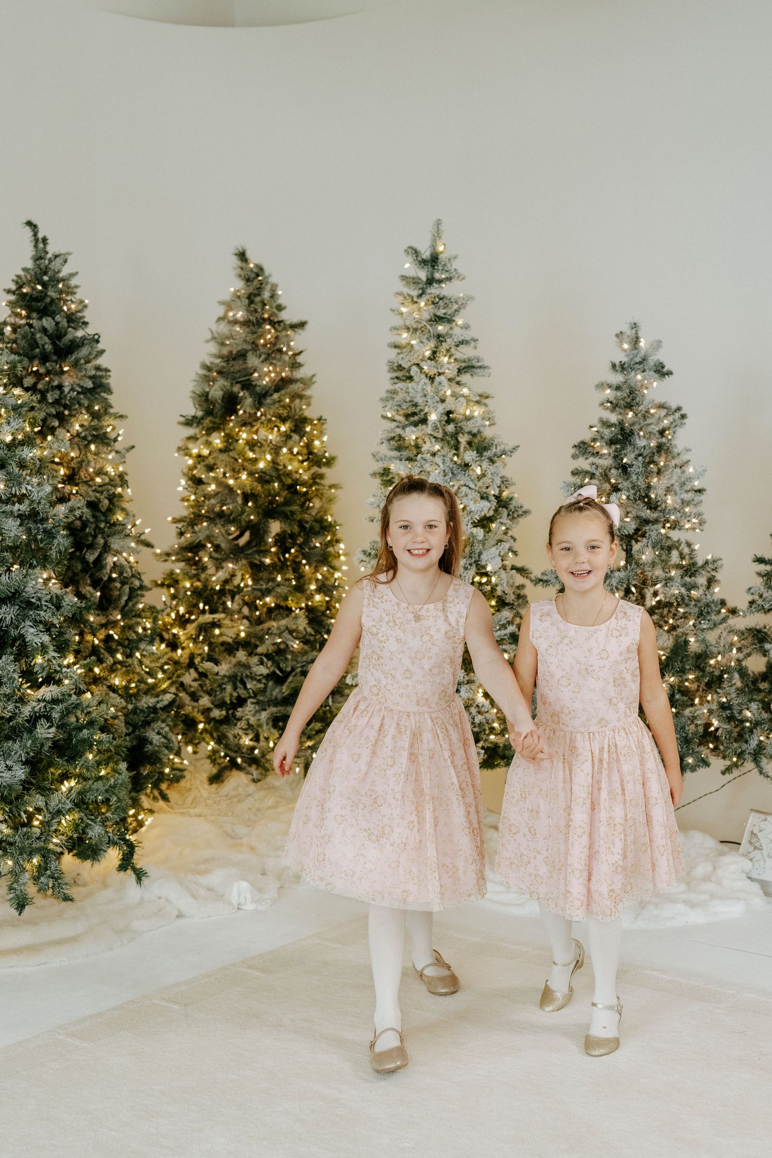Two young children in matching pink sparkly dresses stand together in front of decorated Christmas trees with lights.
