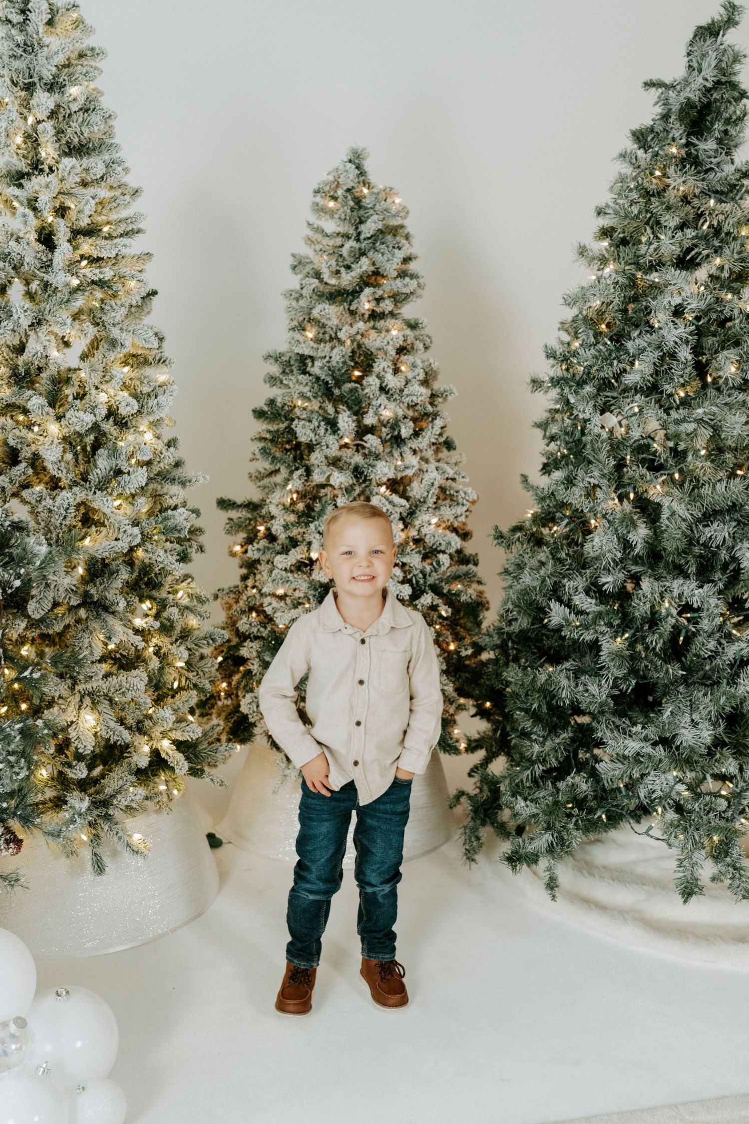 Multiple photos of a child in winter attire posing in front of snow-covered Christmas trees against a white backdrop.