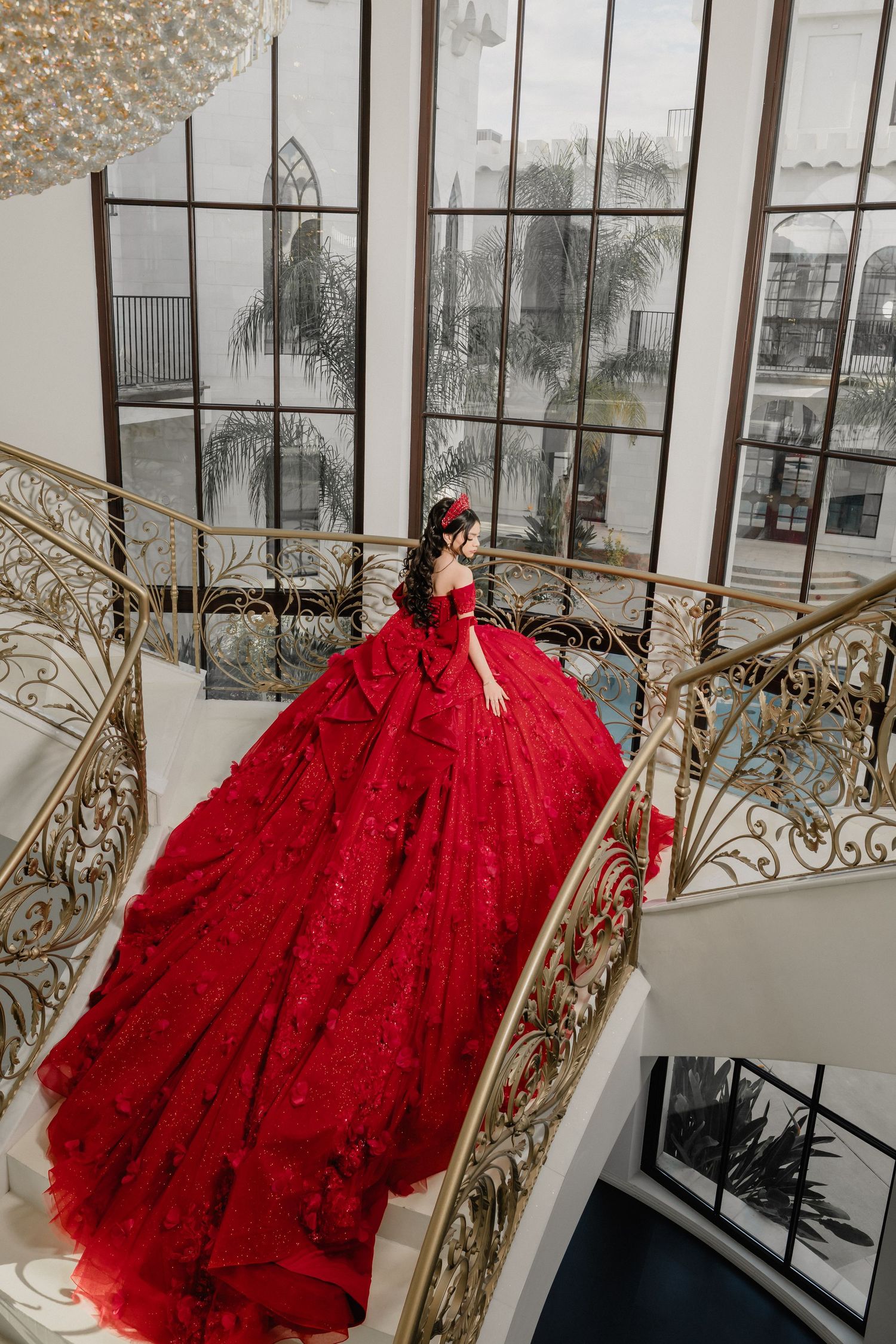 Stunning red ball gown with intricate lace details photographed on an elegant staircase with ornate gold railings.