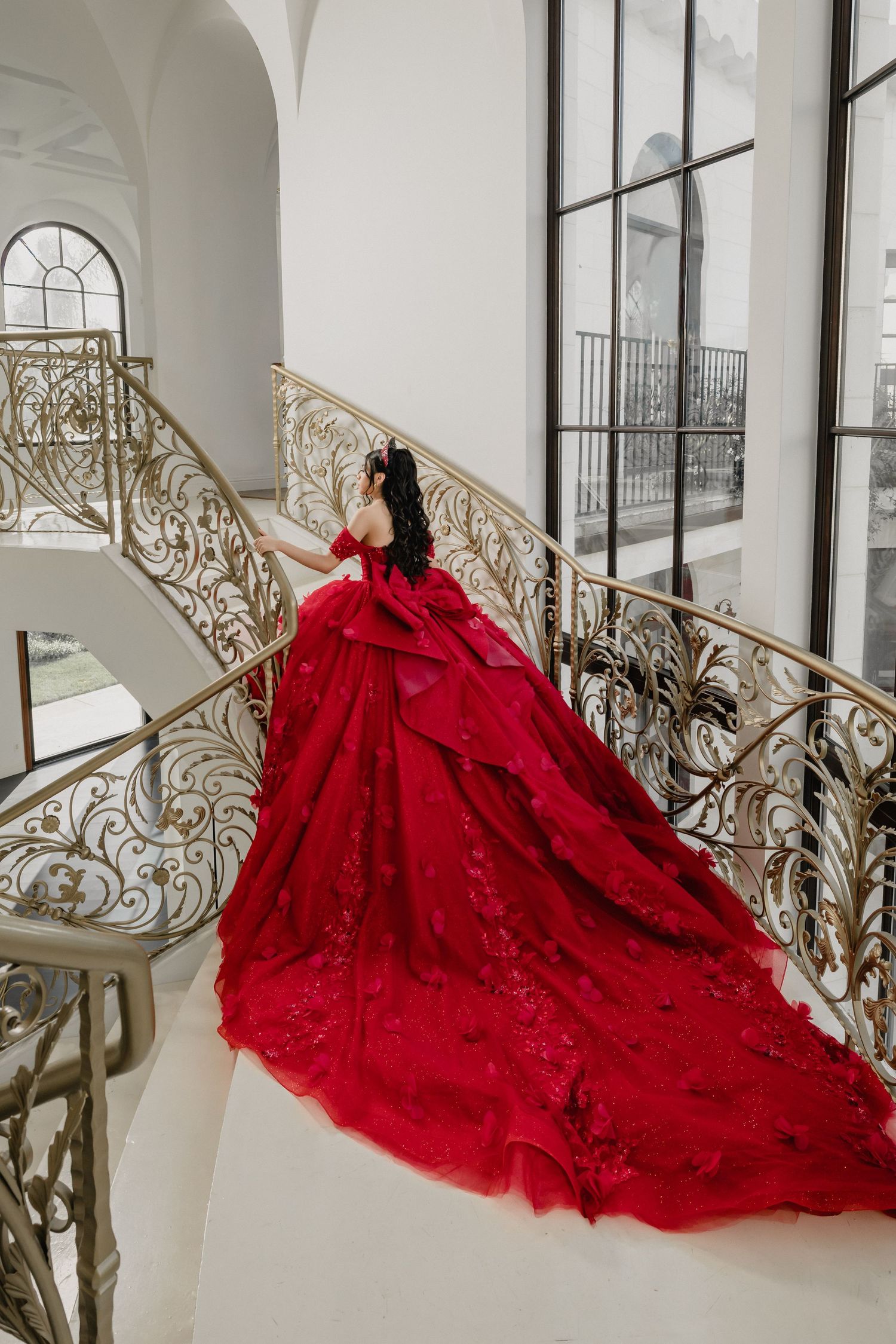 A stunning red ball gown with intricate lace details cascades down an elegant white staircase with ornate gold railings.