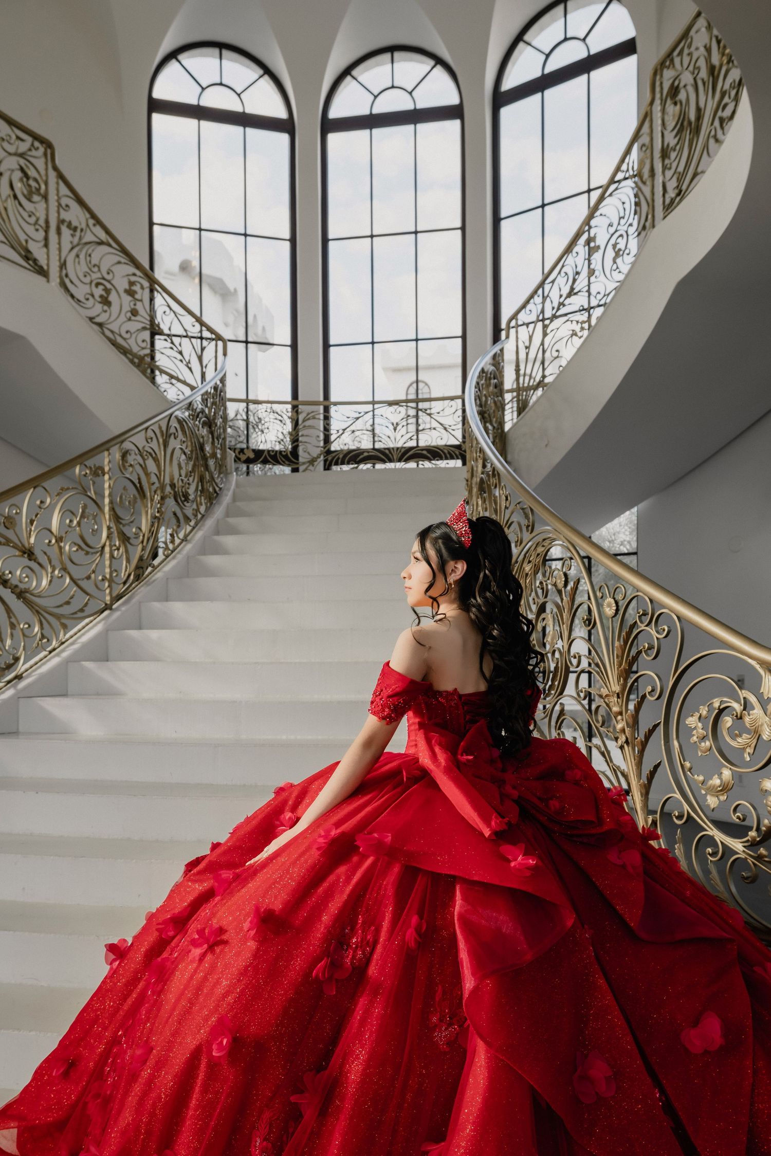 A stunning red ballgown photographed in an elegant staircase setting with arched windows and ornate gold railings.