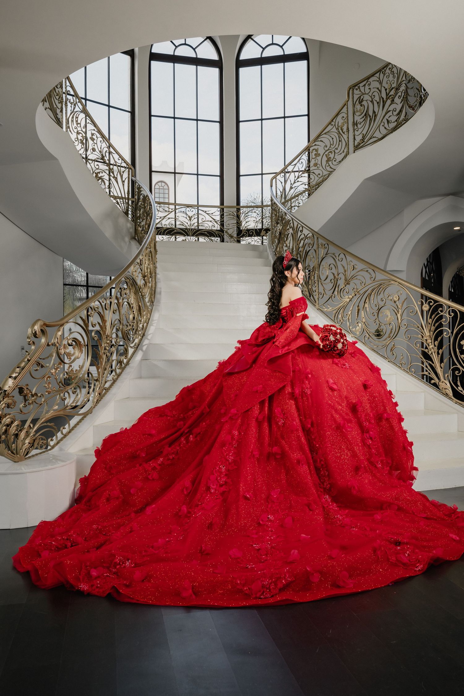 A dramatic red ball gown with a flowing train photographed on a grand spiral staircase with ornate railings.