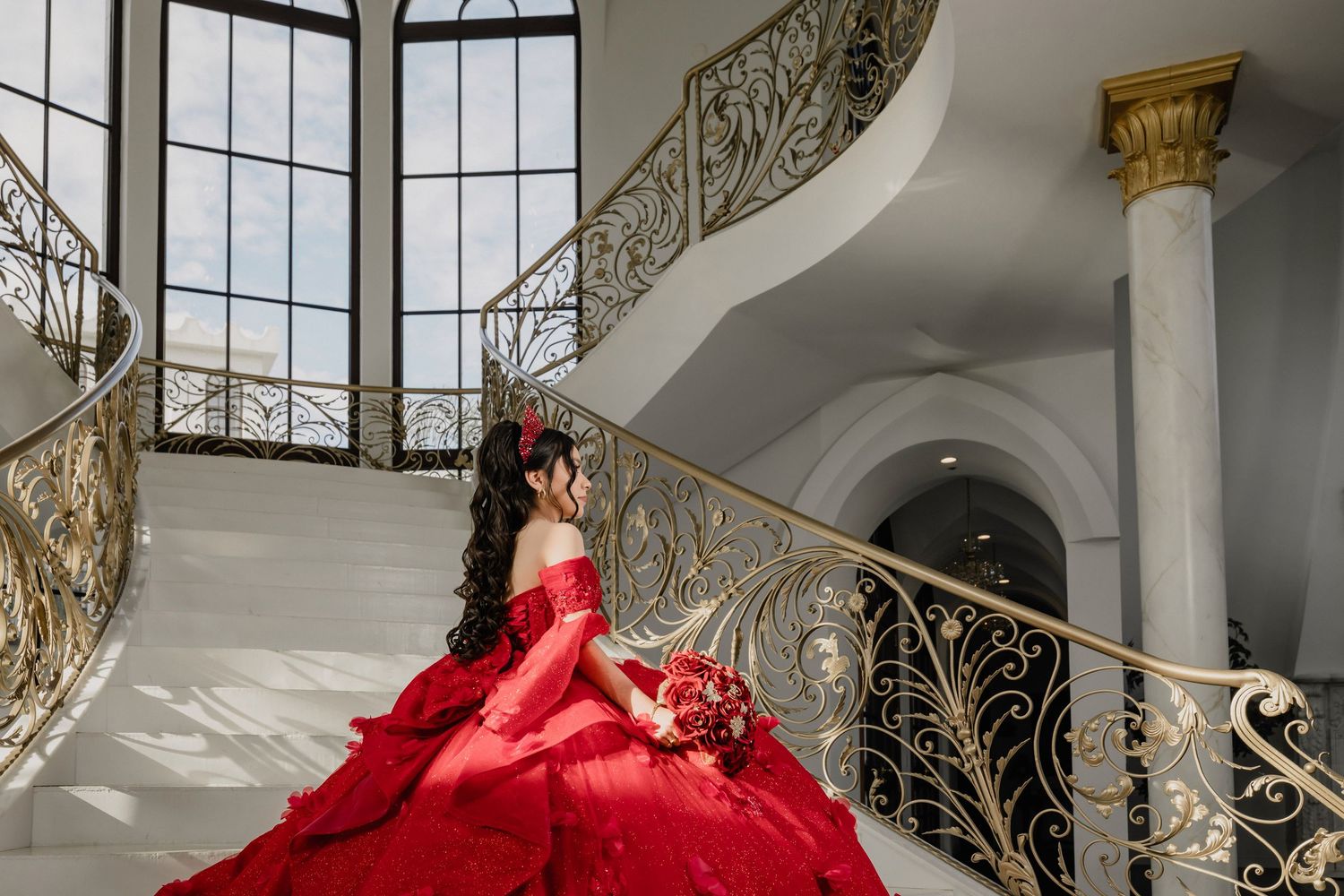 A stunning red ball gown flows down an elegant white marble staircase in a luxurious mansion interior.