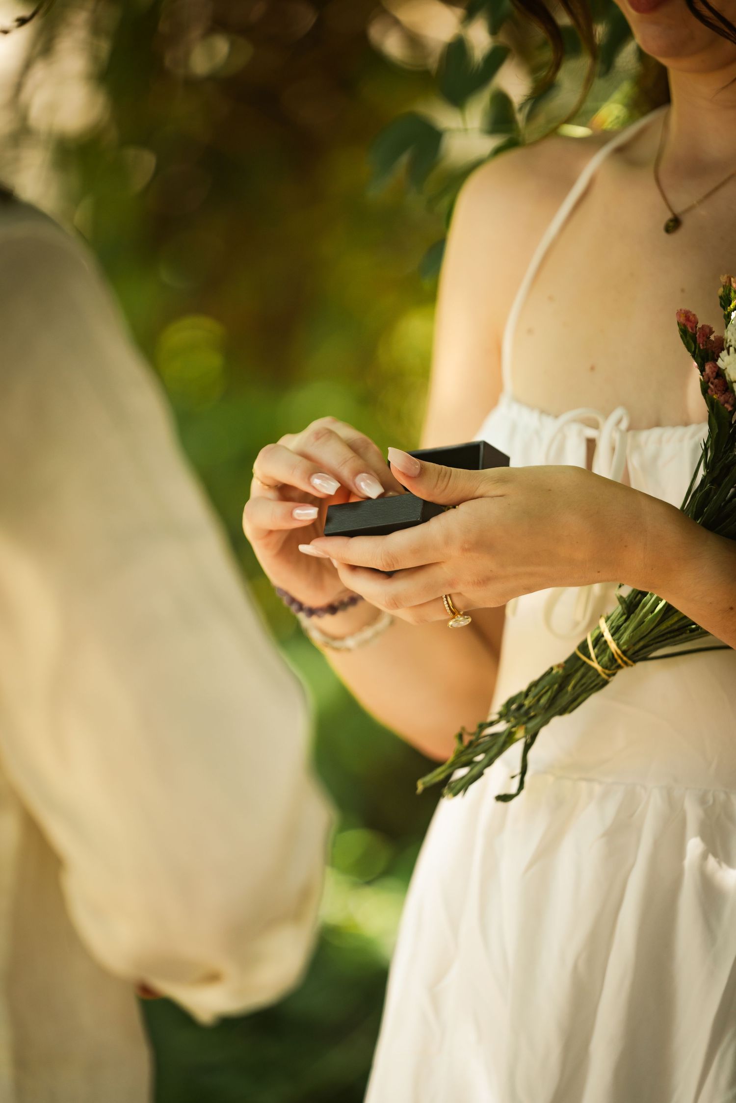 A bride in a white dress holds a ring box while exchanging rings with her partner during an outdoor wedding ceremony.