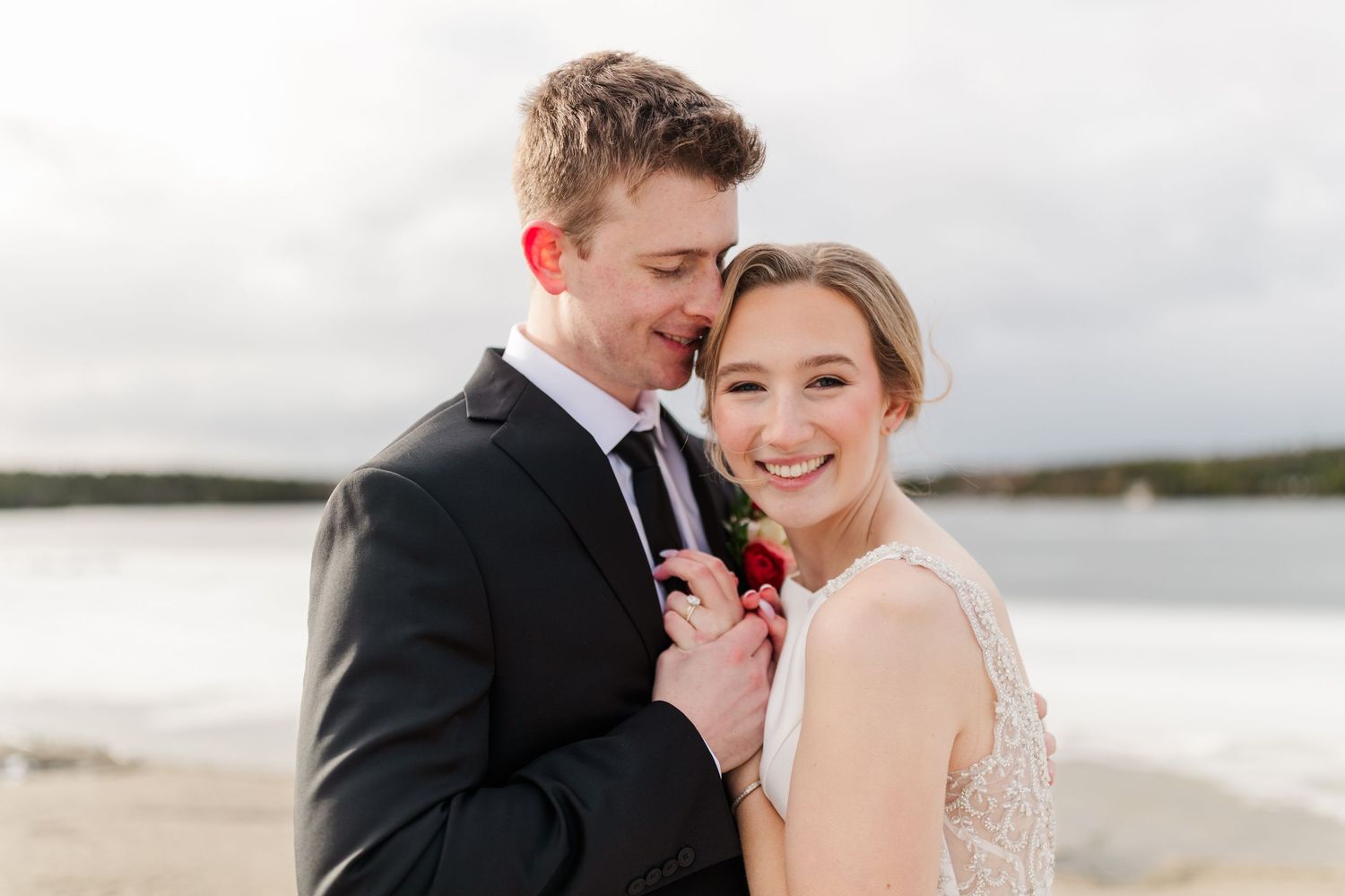 A happy couple embraces on a beach with water and cloudy skies in the background.