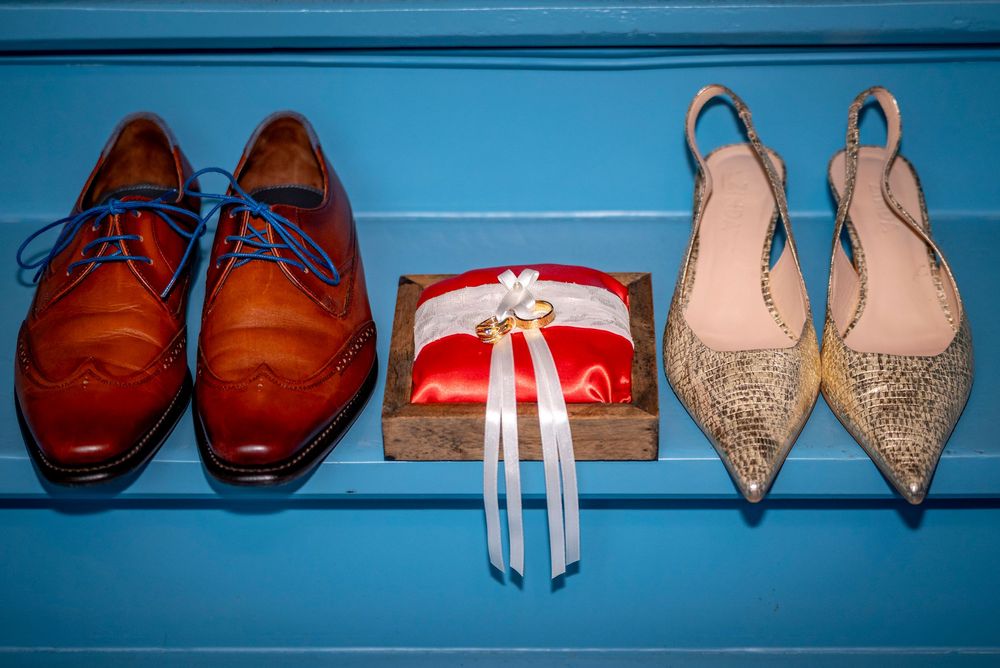 Brown leather brogue shoes, wedding rings on a red pillow, and gold heels arranged on a blue surface.