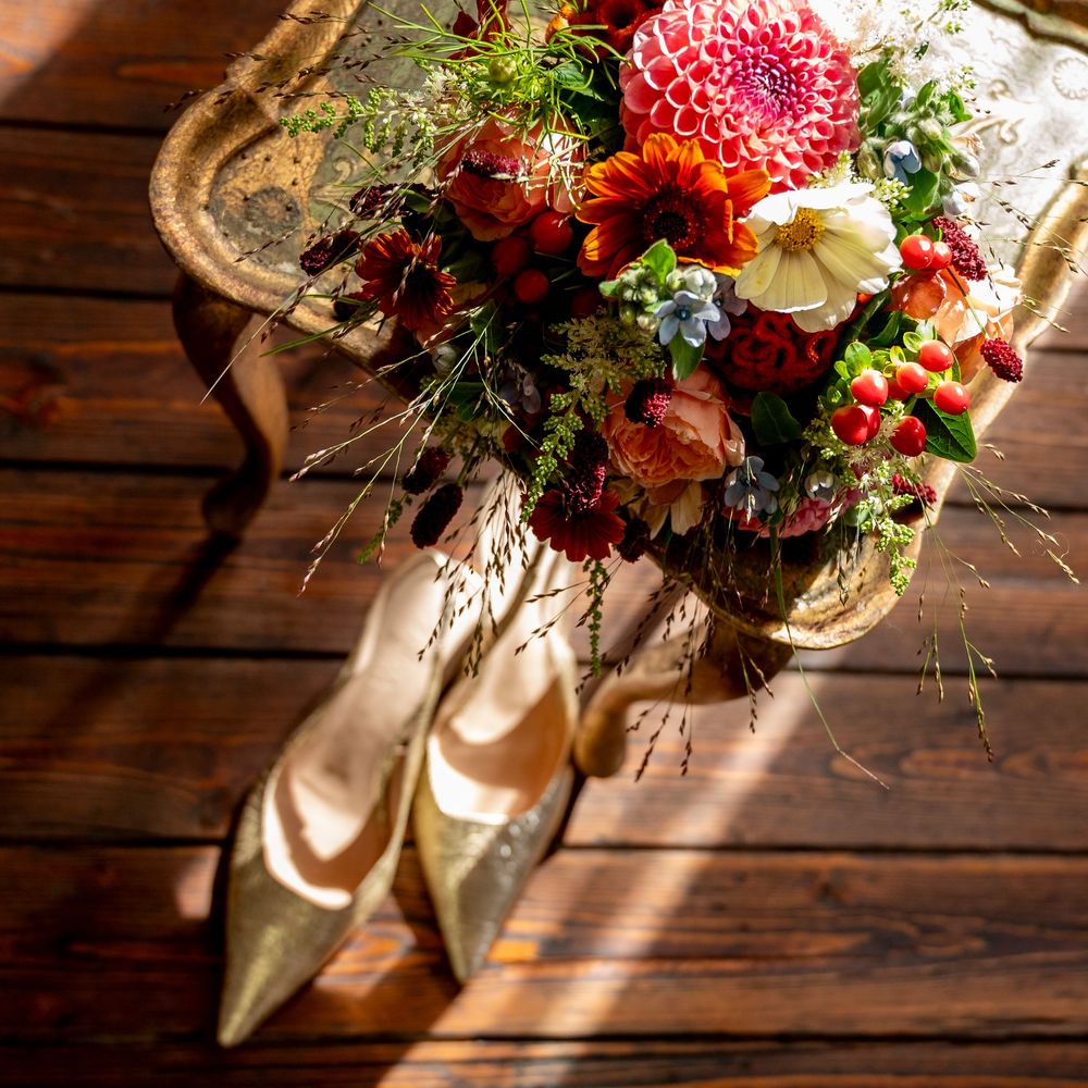 A vibrant autumn bridal bouquet with dahlias and berries beside gold heels on a wooden surface.