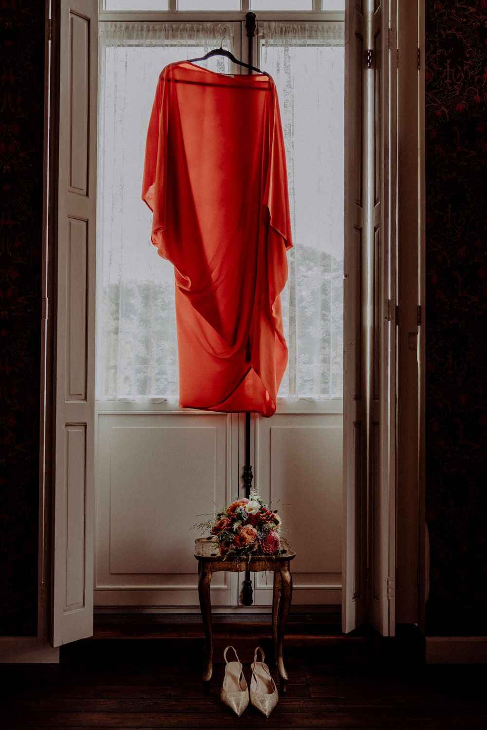 A flowing red dress hangs in a window above white heels and a colorful bridal bouquet.