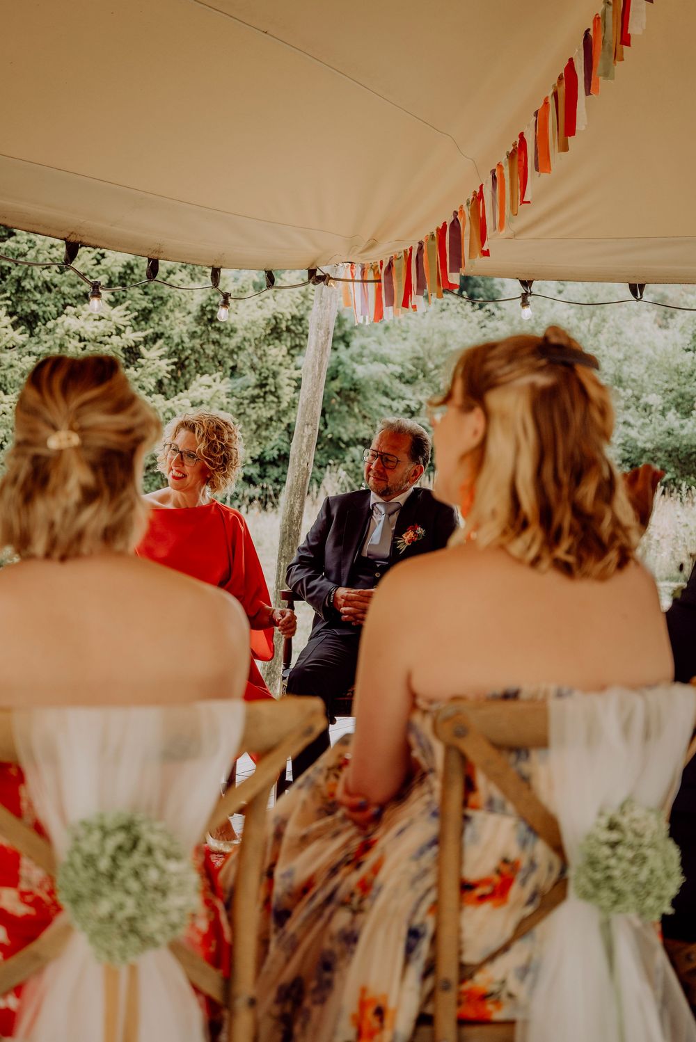 Wedding guests in floral dresses watching couple seated under decorated tent canopy at outdoor ceremony.