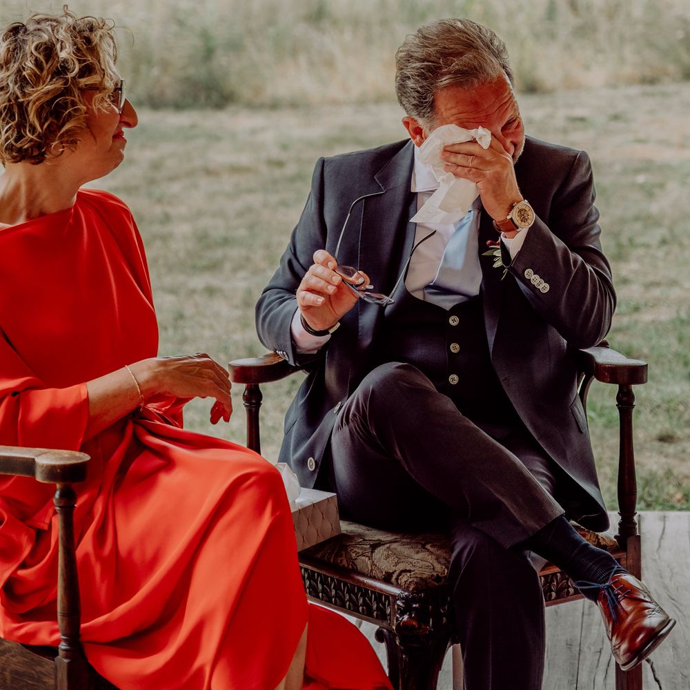 Emotional groom in navy suit wiping tears beside bride in red dress during outdoor wedding ceremony.