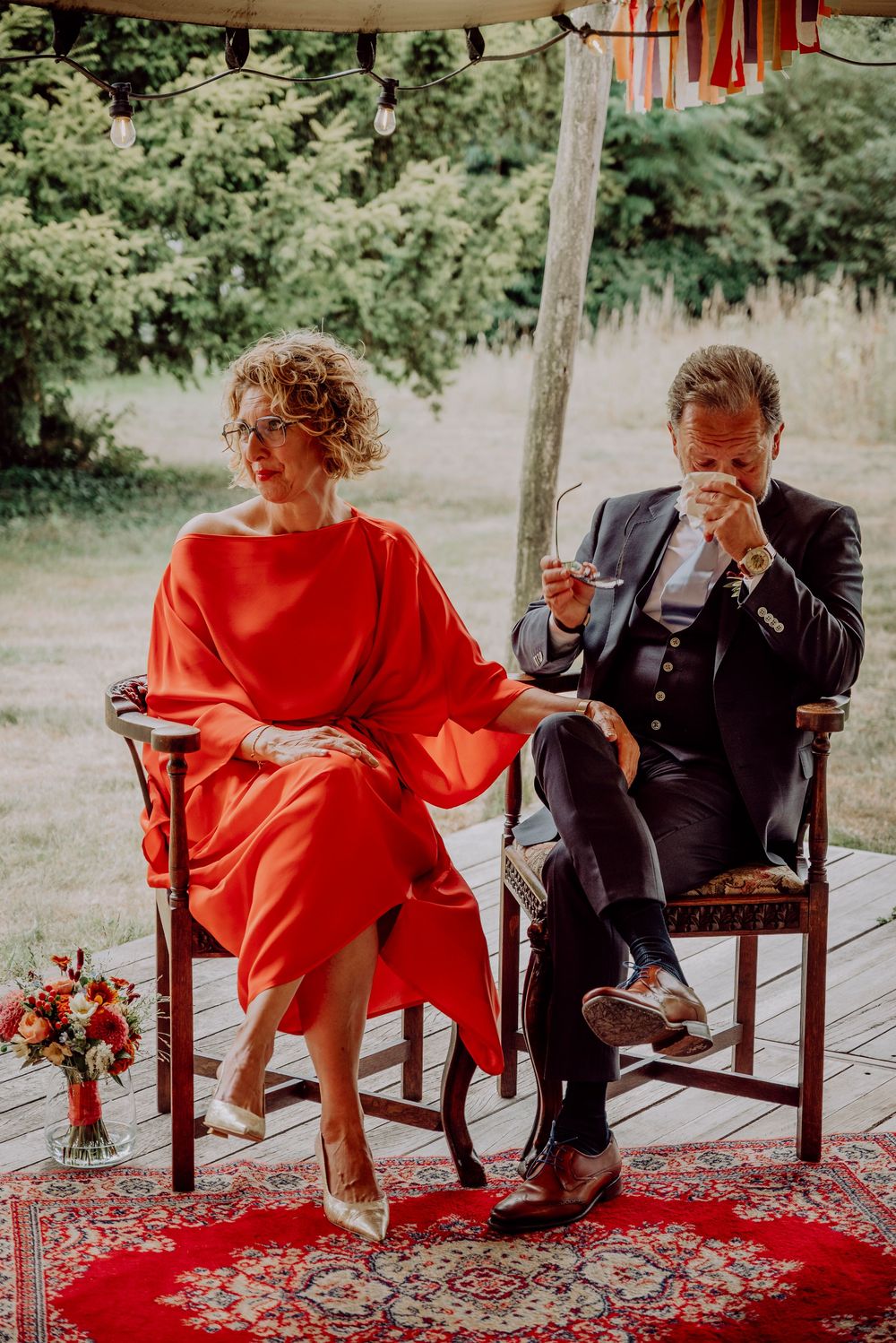 Bride in flowing red dress and groom in navy suit seated together during outdoor wedding ceremony on red rug.
